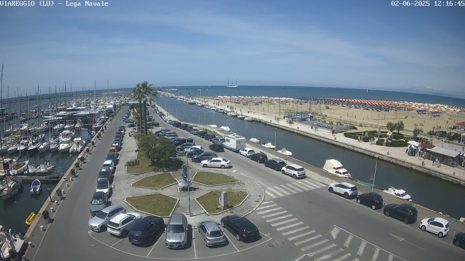 A sunny day overlooking a marina filled with boats, a canal lined with parked cars and a sandy beach with numerous umbrellas.