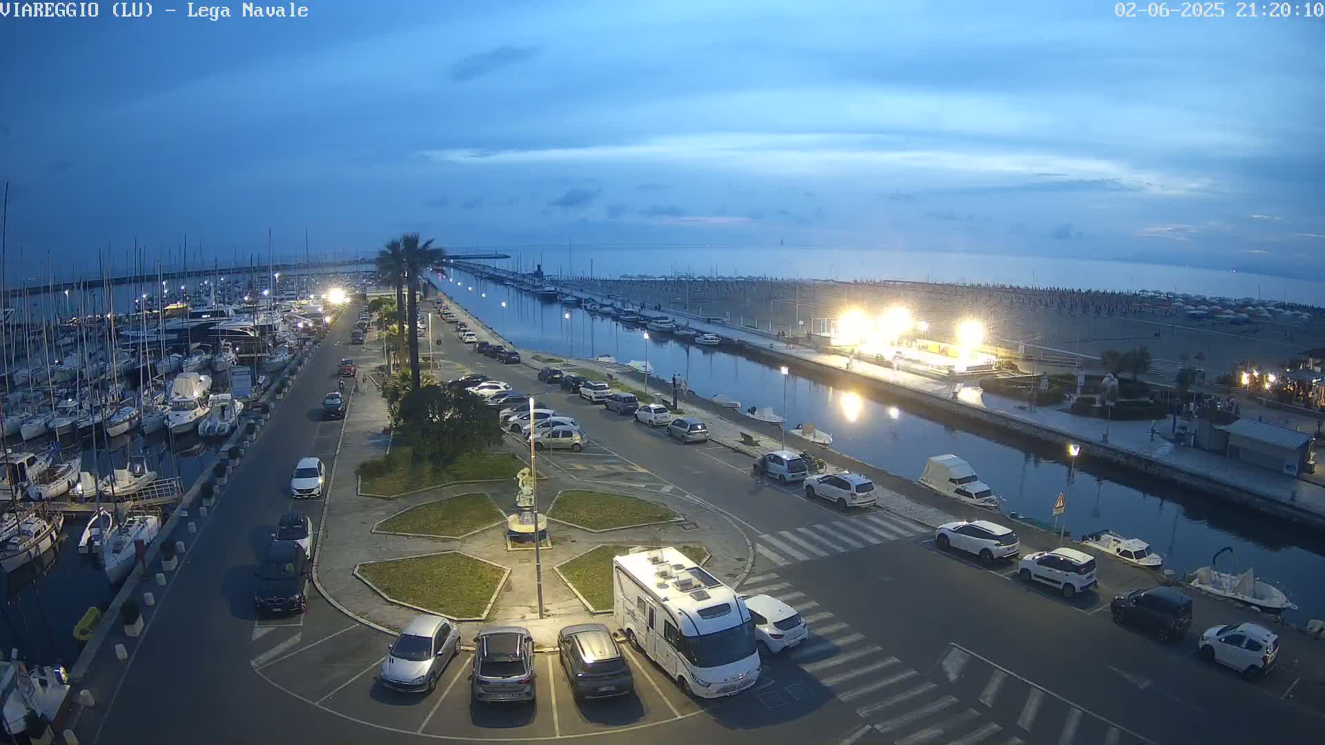 An aerial, twilight view of a marina with numerous boats, a canal, parked cars, and a beach in the background under a clear, dusky sky.