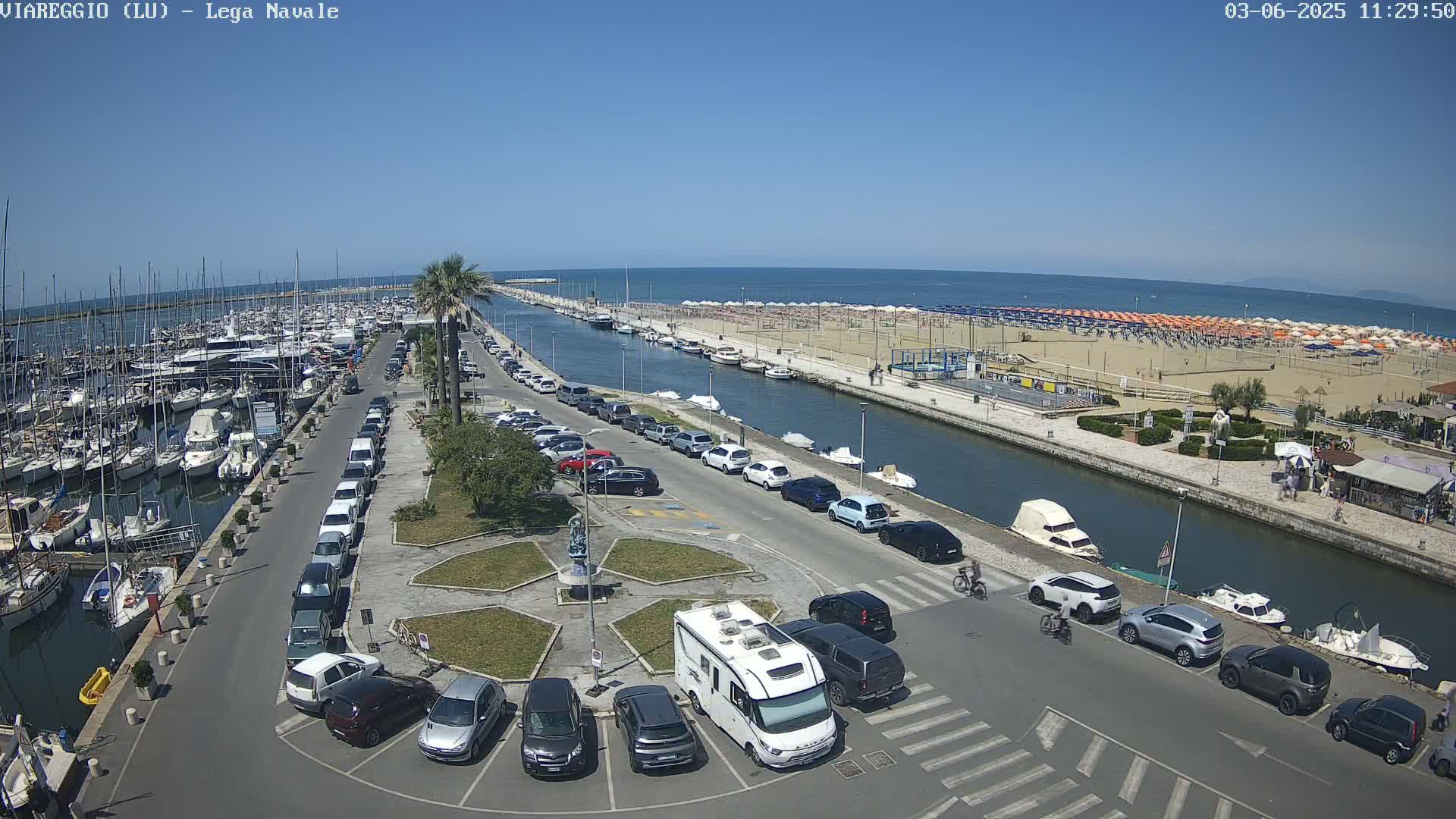 A marina filled with boats, adjacent to a sandy beach with numerous umbrellas under a clear blue sky, is bordered by a canal lined with parked cars and motorhomes.