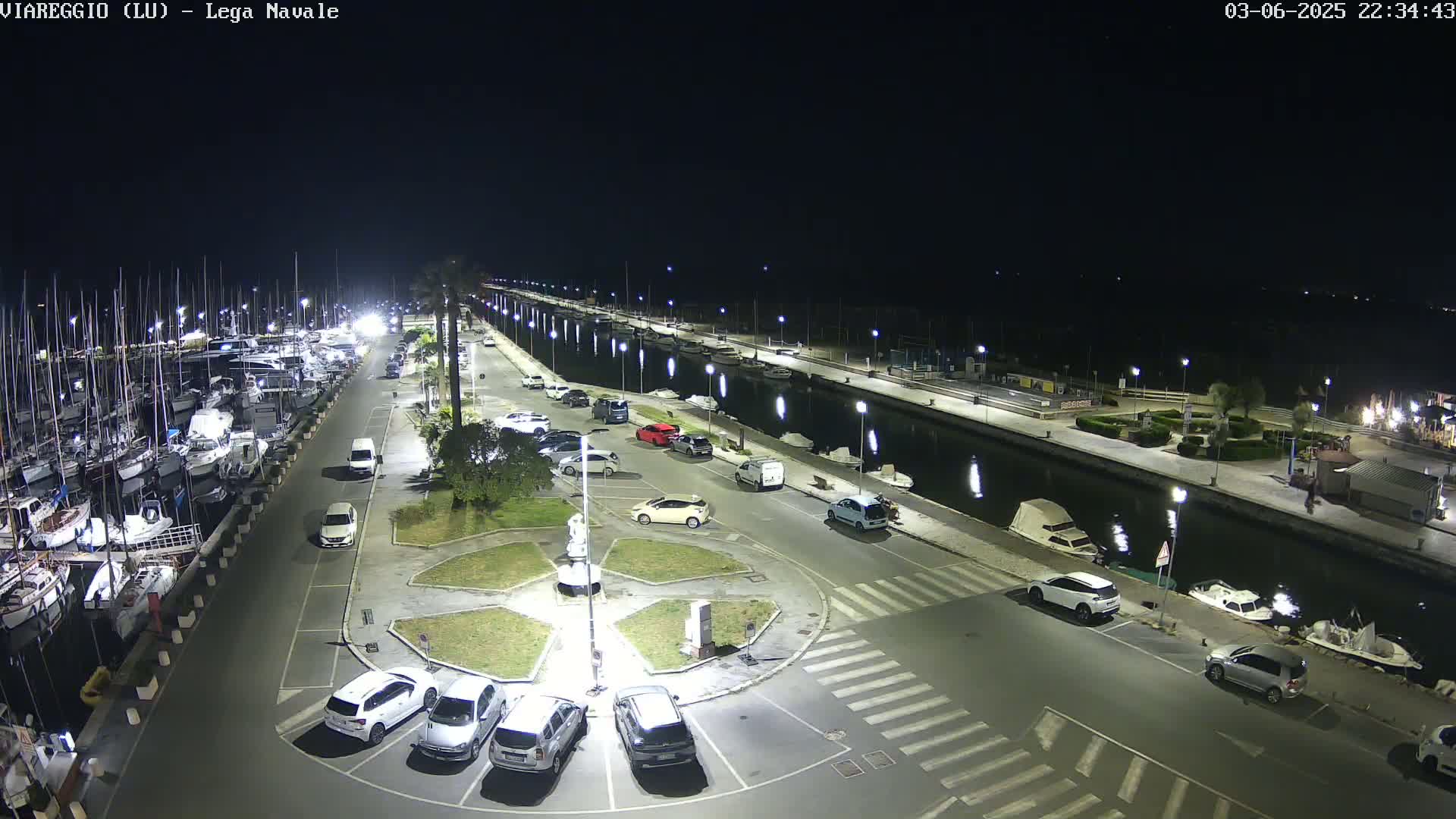 A nighttime aerial view of a marina with numerous boats docked, cars parked in a lot adjacent to a canal, and a statue in a circular plaza.