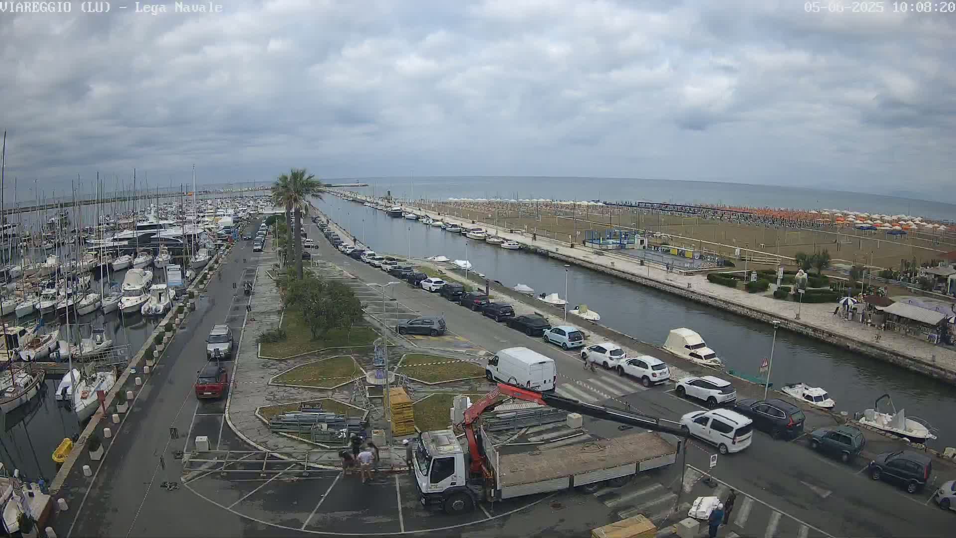 A harbor filled with boats and a sandy beach are visible under a cloudy sky, with cars parked along a canal separating them.