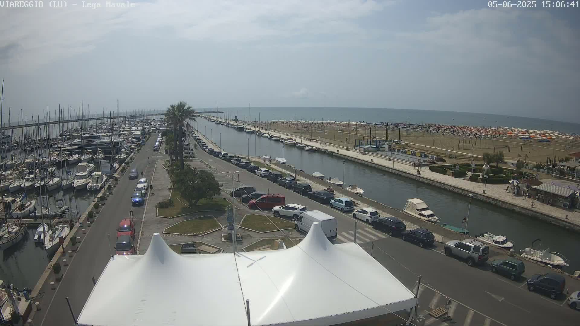 A marina filled with boats is next to a canal that runs alongside a beach with many umbrellas under a partly cloudy sky.