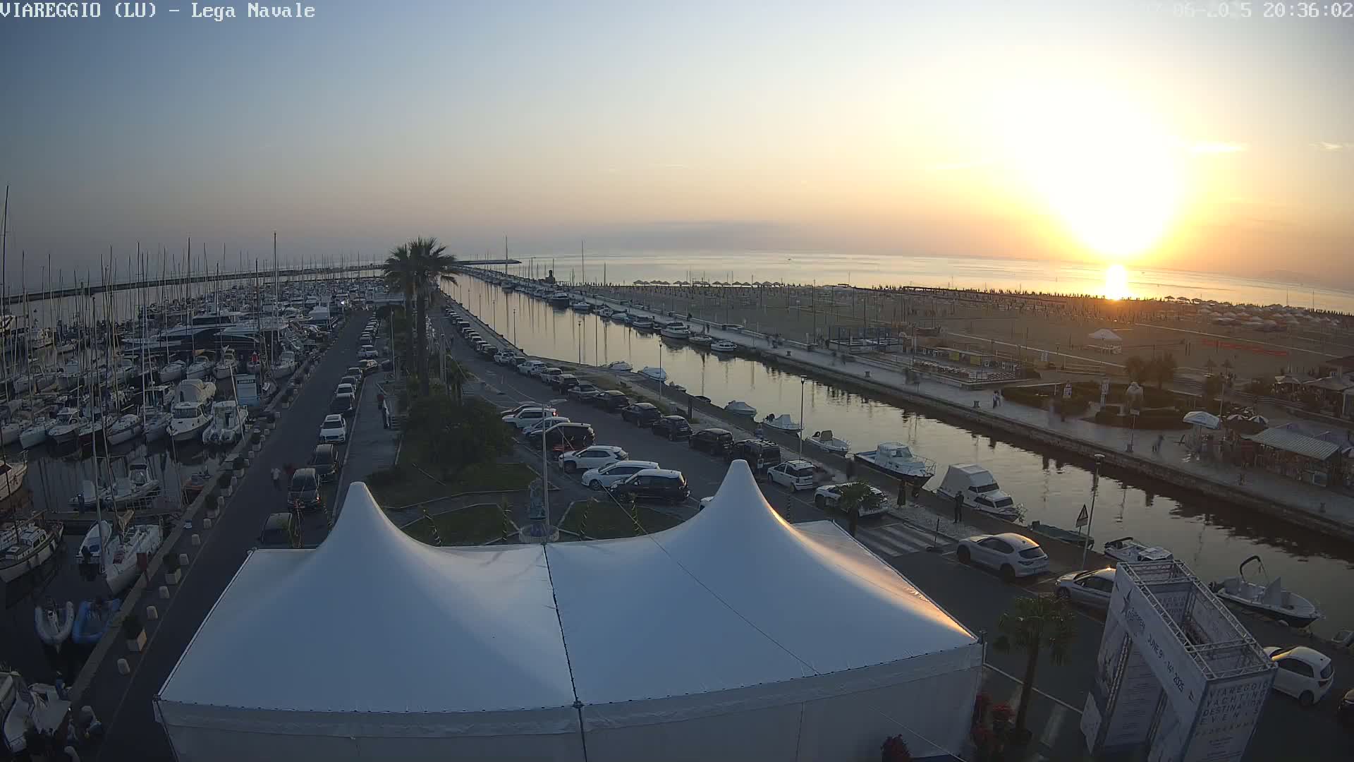 A marina filled with boats at sunset, next to a canal and beach with parked cars and a large white tent in the foreground.