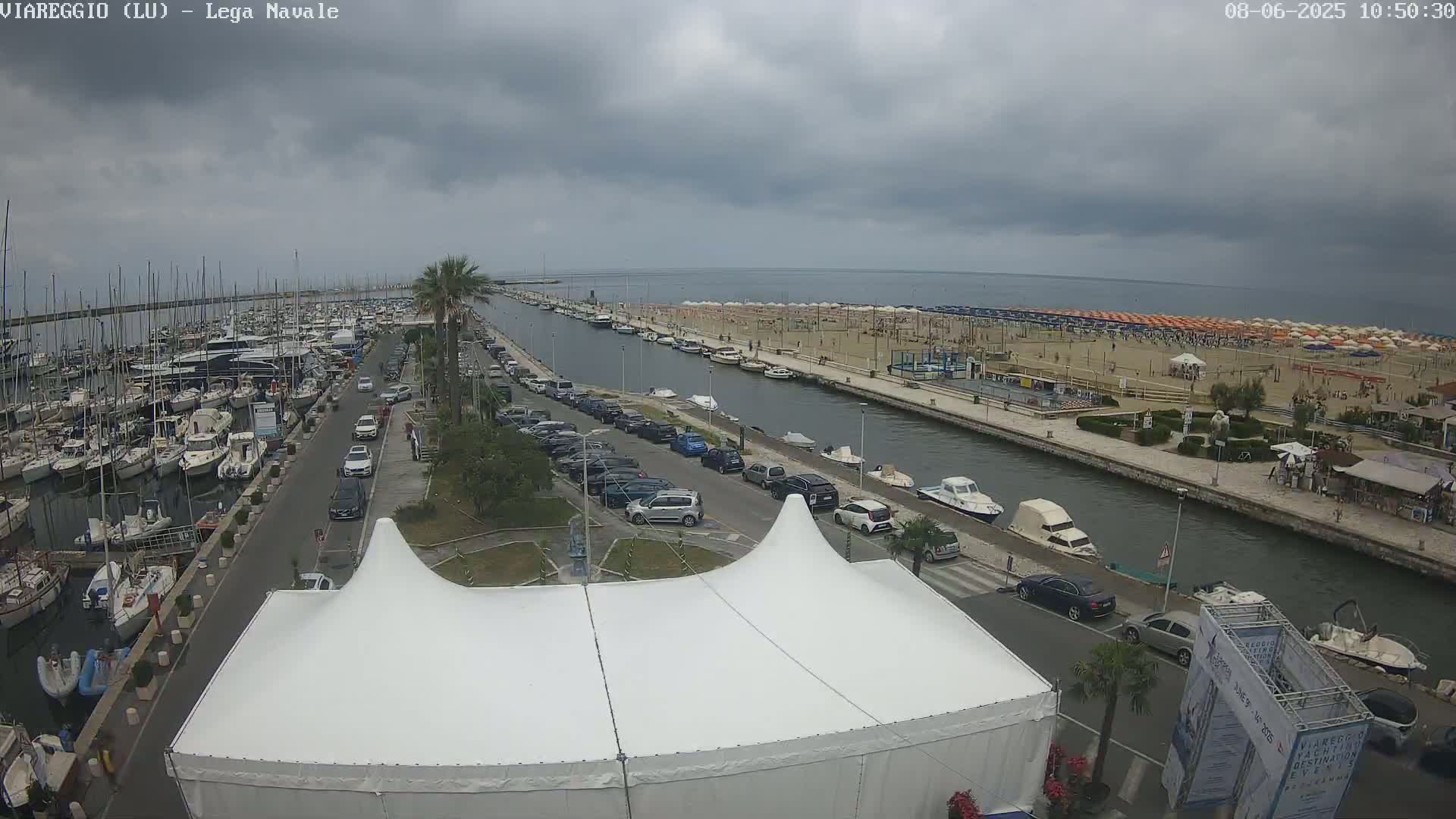 A marina filled with boats is adjacent to a canal that leads to a beach with many umbrellas under an overcast sky.