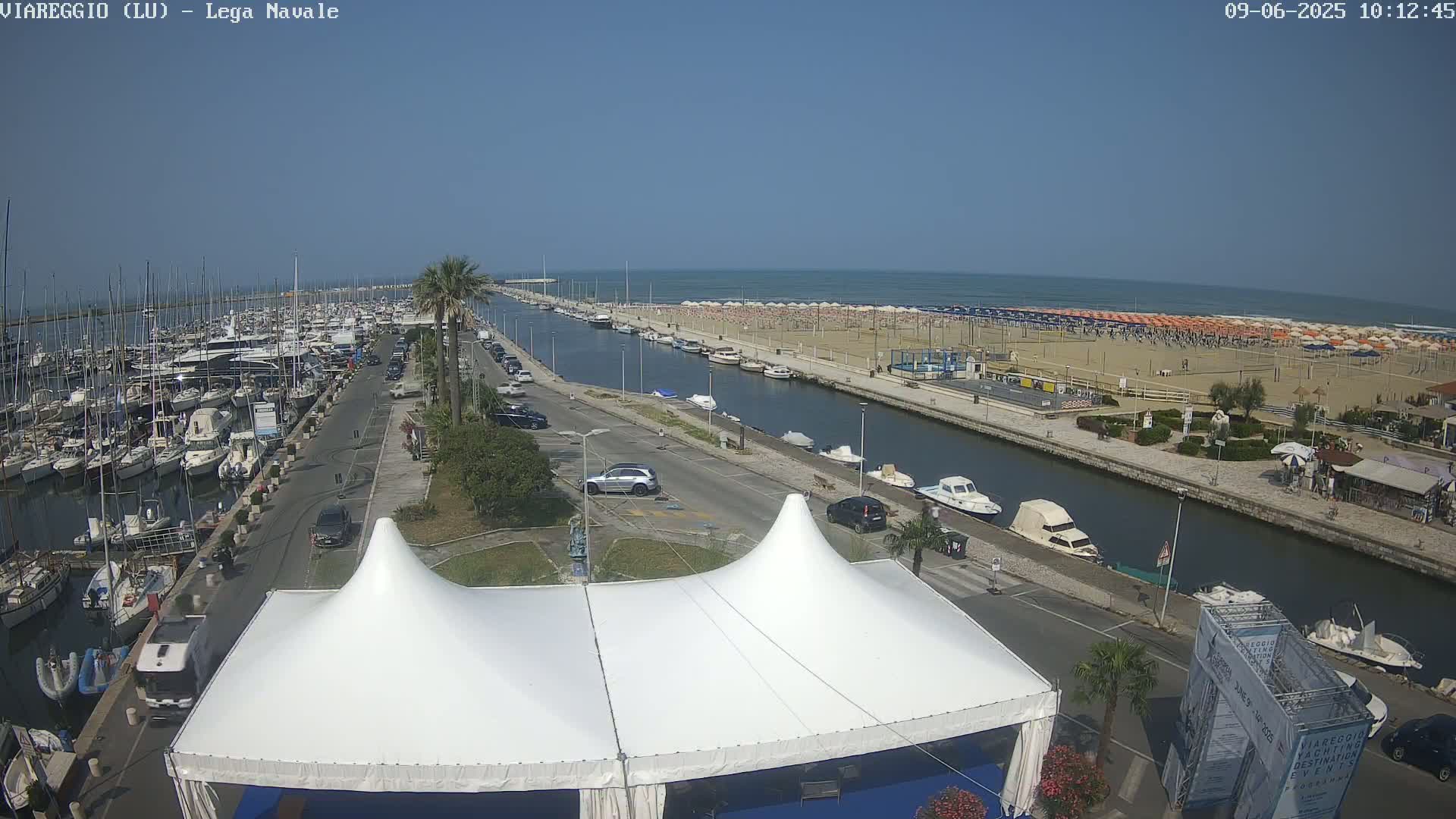 A marina filled with boats is adjacent to a canal that runs alongside a beach crowded with umbrellas and people under a clear, sunny sky.