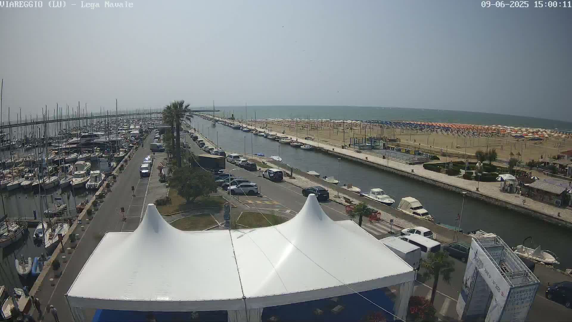 A marina filled with boats is adjacent to a canal that leads to a sandy beach with numerous umbrellas under a clear, sunny sky.