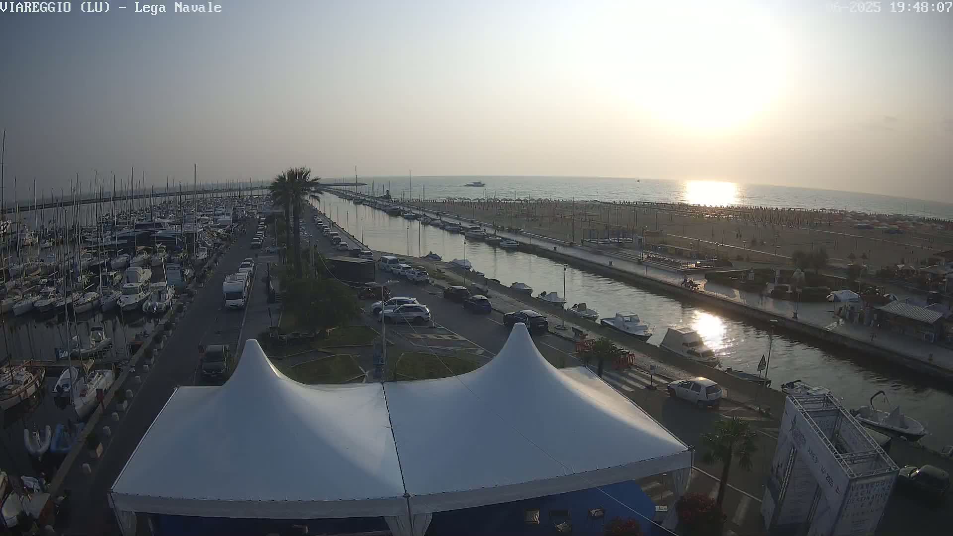 A harbor filled with boats, a canal, and a beach with people are seen at sunset under a mostly clear sky.