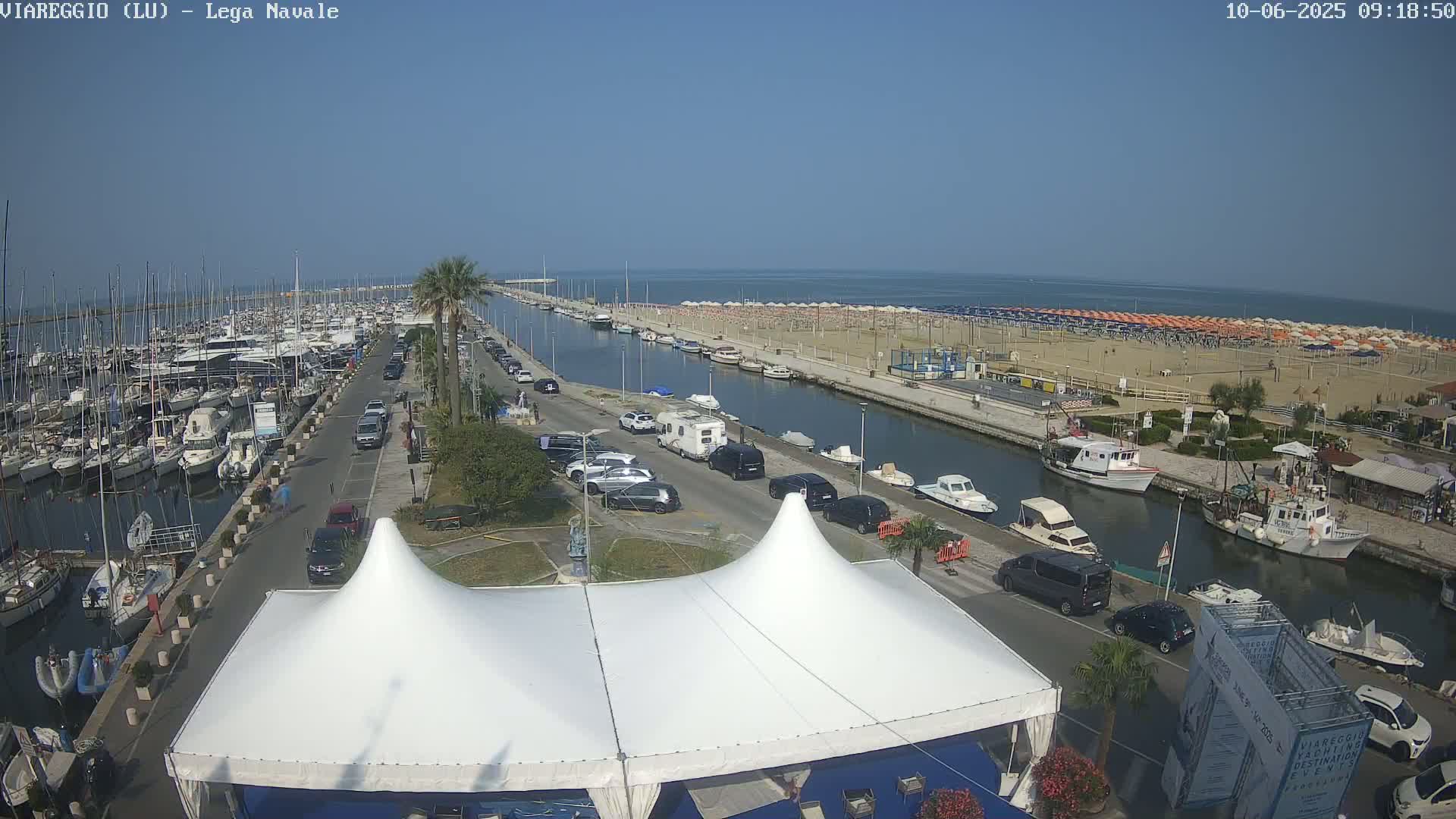 A marina filled with boats is adjacent to a sandy beach with numerous umbrellas under a clear blue sky.