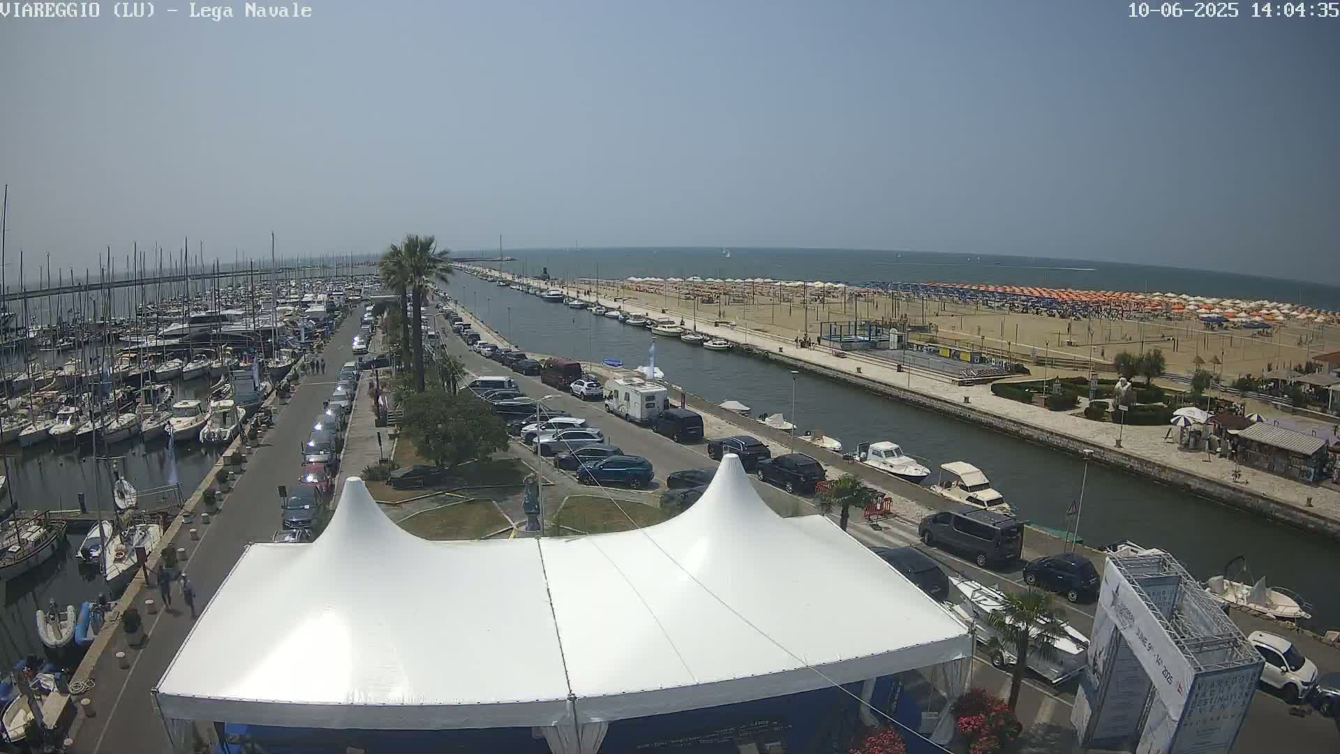 A marina filled with boats, a canal, a sandy beach with numerous umbrellas, and a road lined with parked cars are visible on a sunny day.