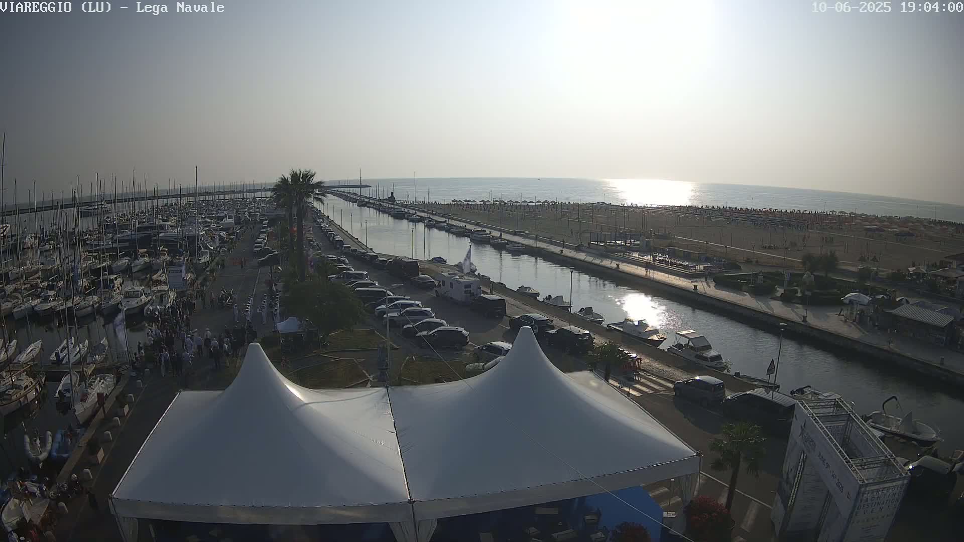 A harbor filled with boats under a mostly sunny sky, with a canal separating it from a beach crowded with people.