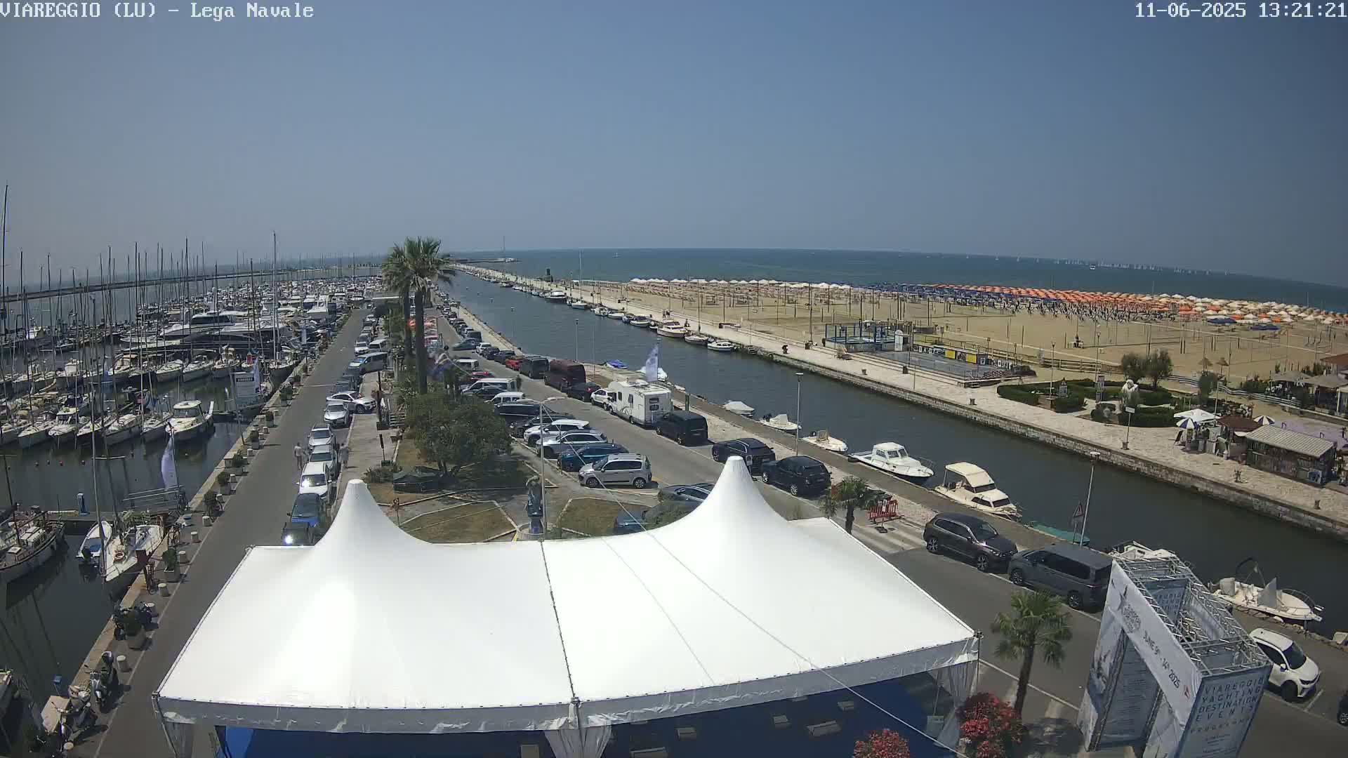 A marina filled with boats is next to a canal that runs alongside a beach with many umbrellas under a clear, sunny sky.