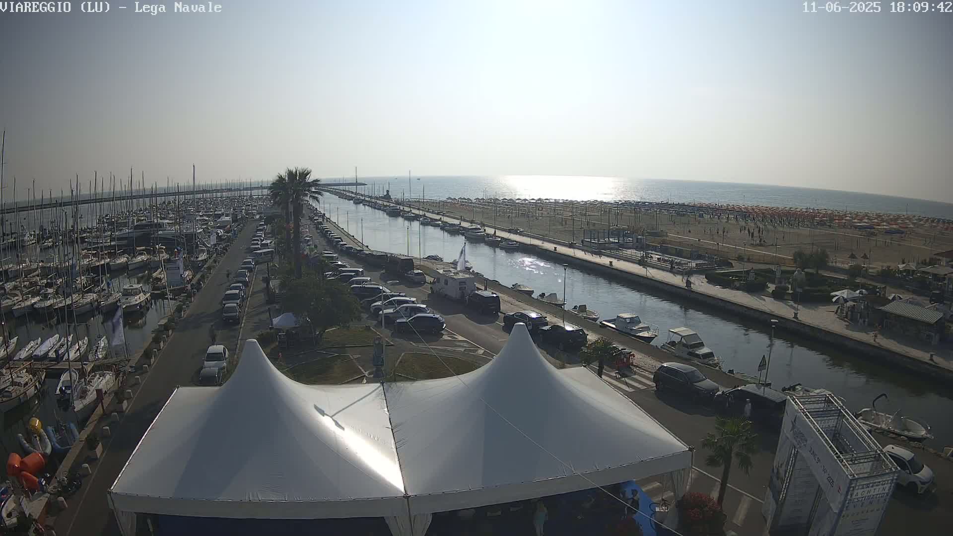 A sunny day overlooking a harbor filled with boats, a canal, a beach with many umbrellas, and a street with parked cars and a large white tent.