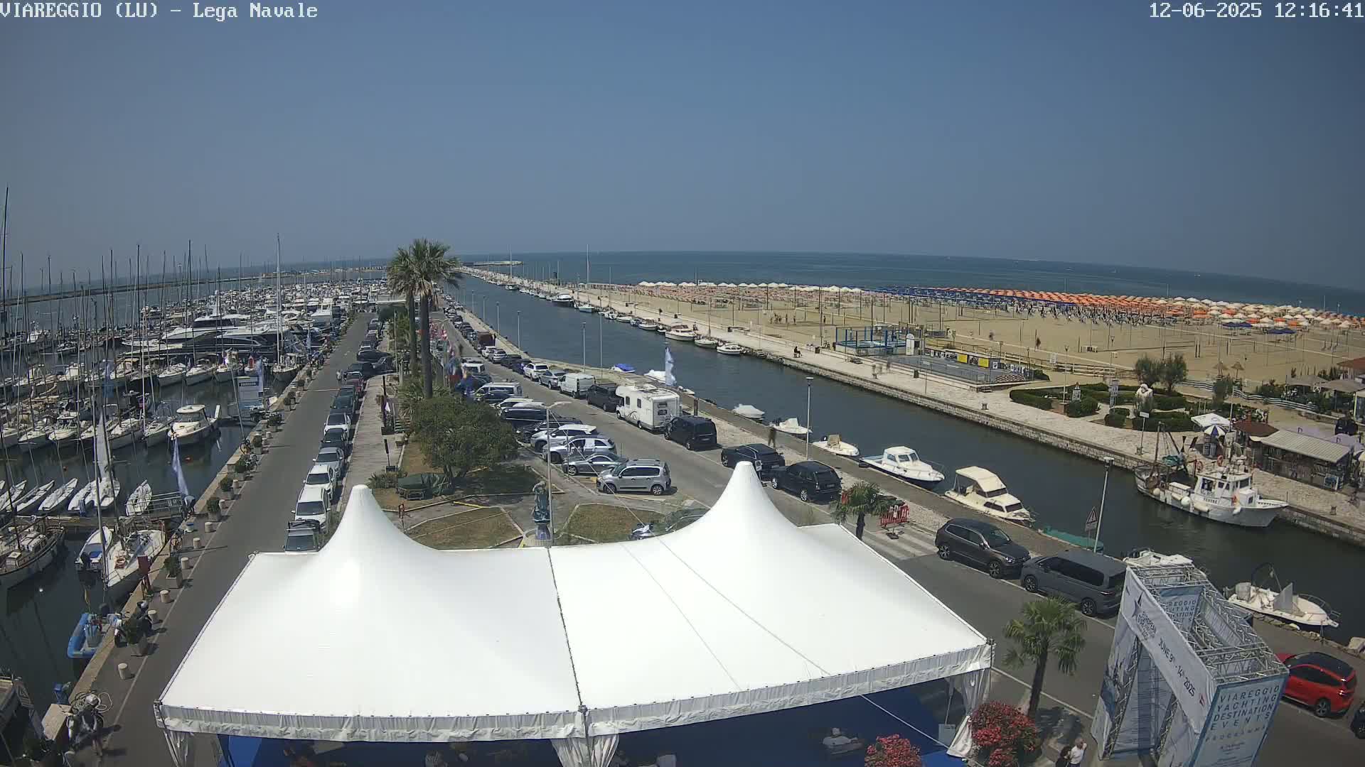 A marina filled with boats, a sandy beach lined with umbrellas, and a road with parked cars are seen on a sunny day.