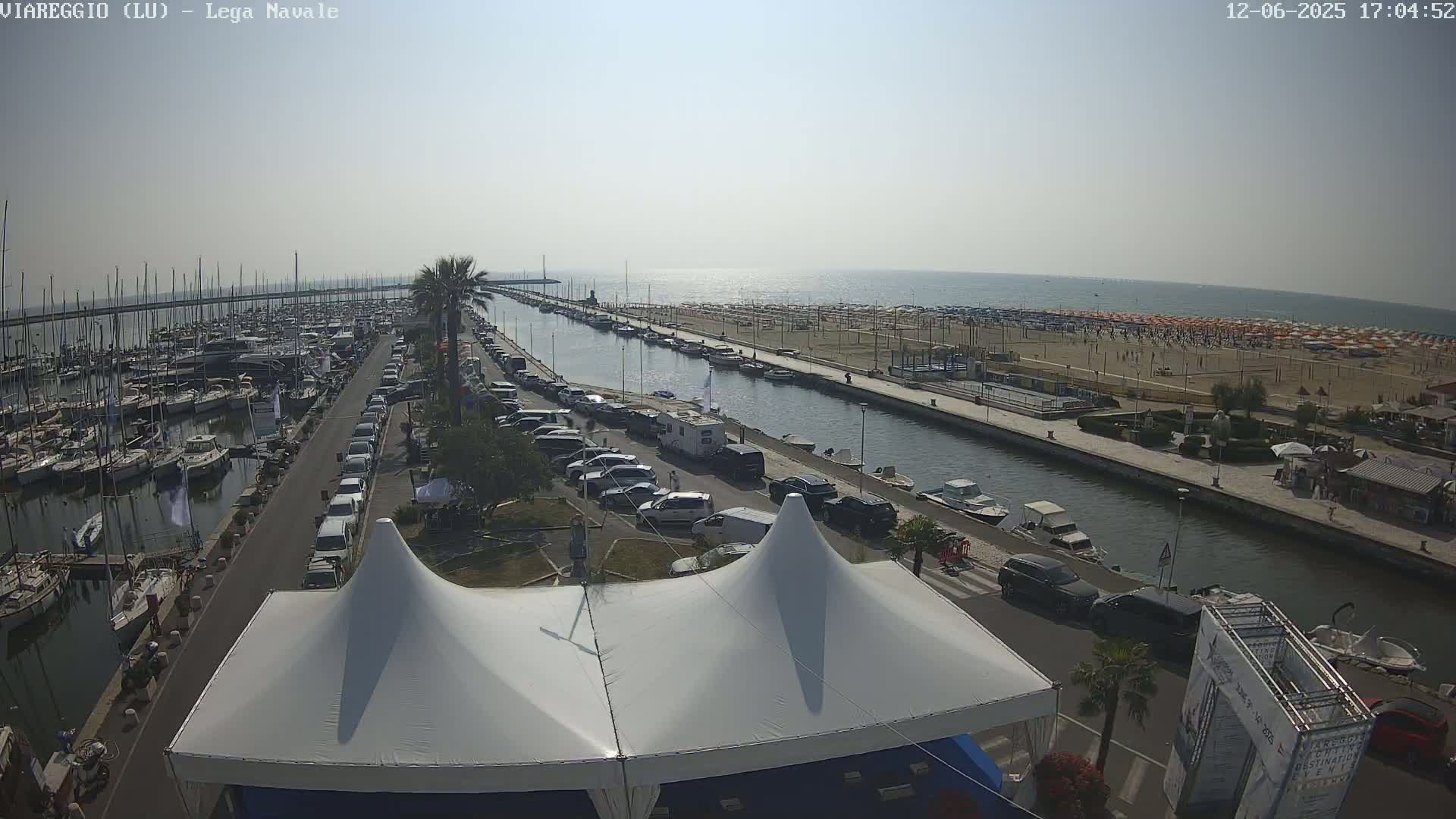 A marina filled with boats is next to a canal that leads to a sandy beach with numerous umbrellas under a clear, sunny sky.