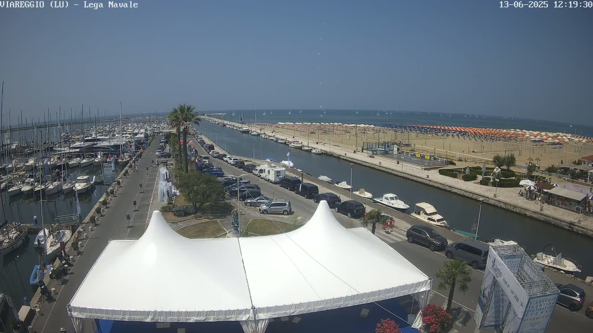 A sunny day at a harbor shows many boats docked, a canal with cars parked alongside, a beach with numerous umbrellas, and a large white tent.
