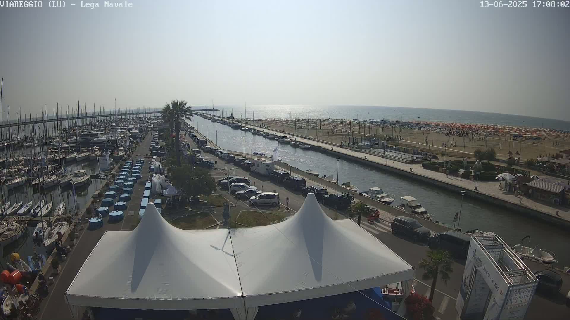 A sunny day at a harbor shows many boats docked, a canal with parked cars along its side, a sandy beach with numerous umbrellas, and a large white tent in the foreground.