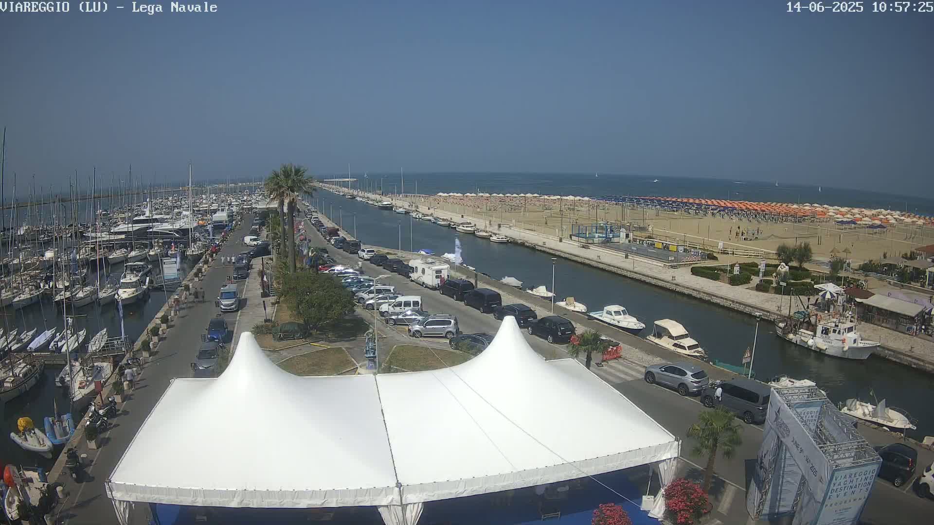 A harbor filled with boats, a sandy beach with numerous umbrellas, and a street lined with parked cars are visible under a clear blue sky.