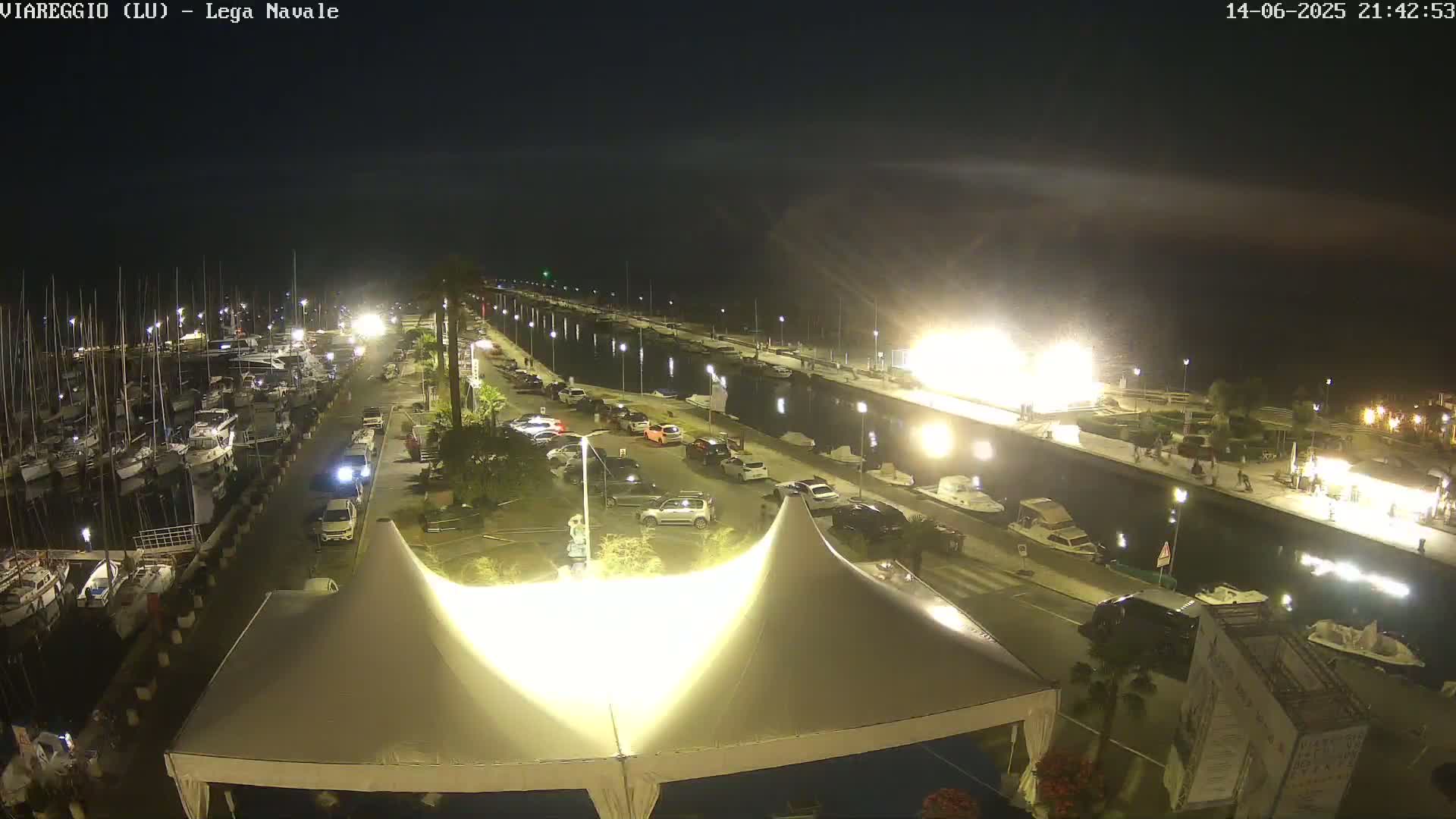 A marina filled with boats is next to a canal and a beach covered in umbrellas under a clear sky.