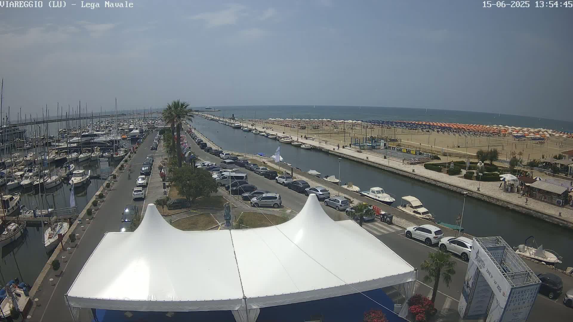 A sunny day shows a marina filled with boats, a canal lined with parked cars and beach umbrellas on a sandy beach beyond.