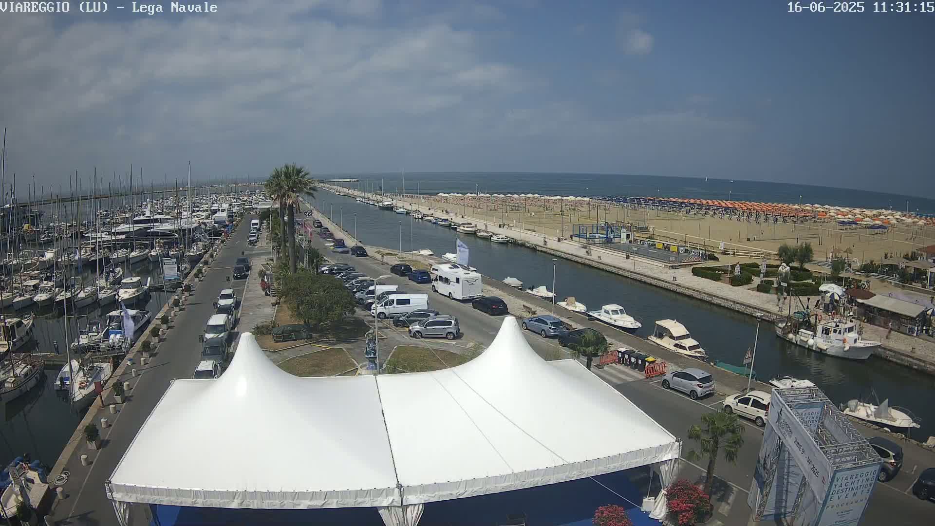 A marina filled with boats is adjacent to a beach with numerous umbrellas under a clear, sunny sky, all viewed from above a white tent.