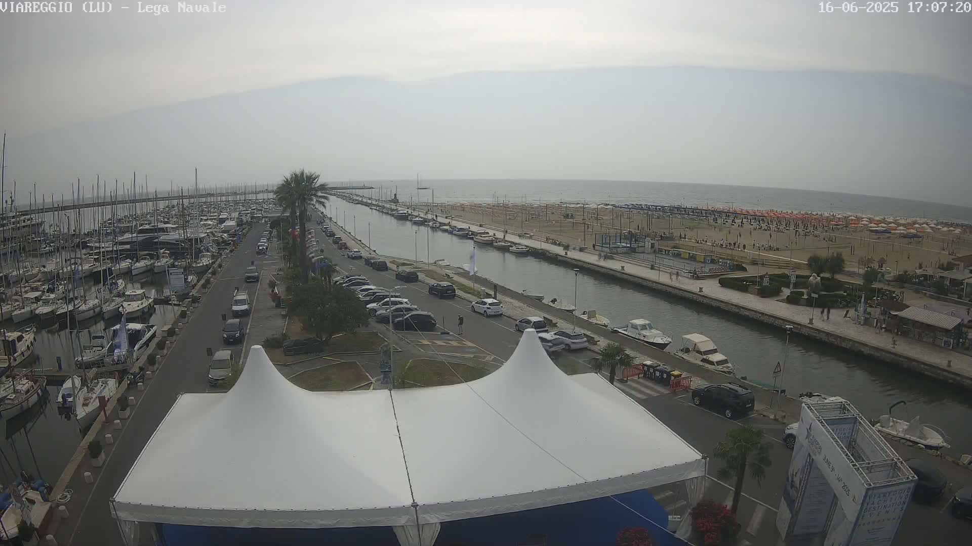 A marina filled with boats, a canal, a beach with numerous umbrellas, and a street with parked cars are visible under an overcast sky.