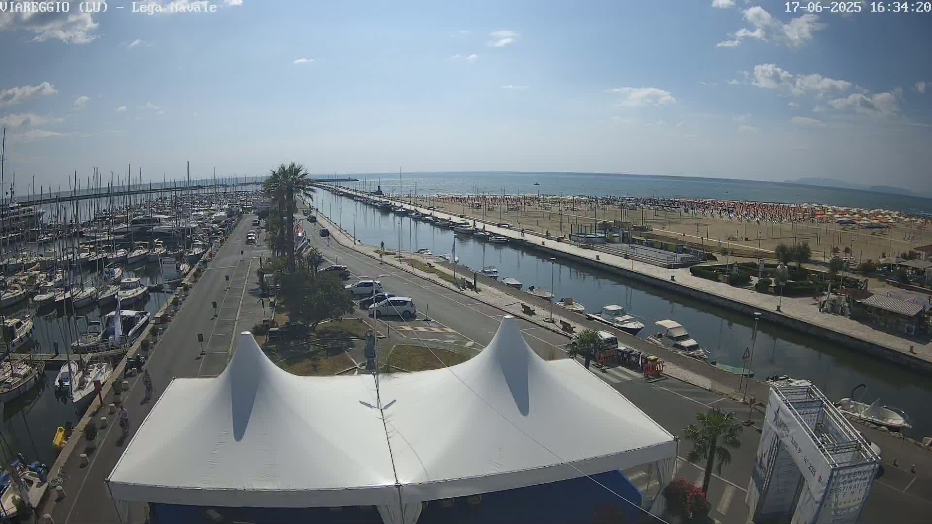 A marina filled with boats, a canal, a beach with numerous umbrellas, and a street with parked cars are seen under a partly sunny sky.