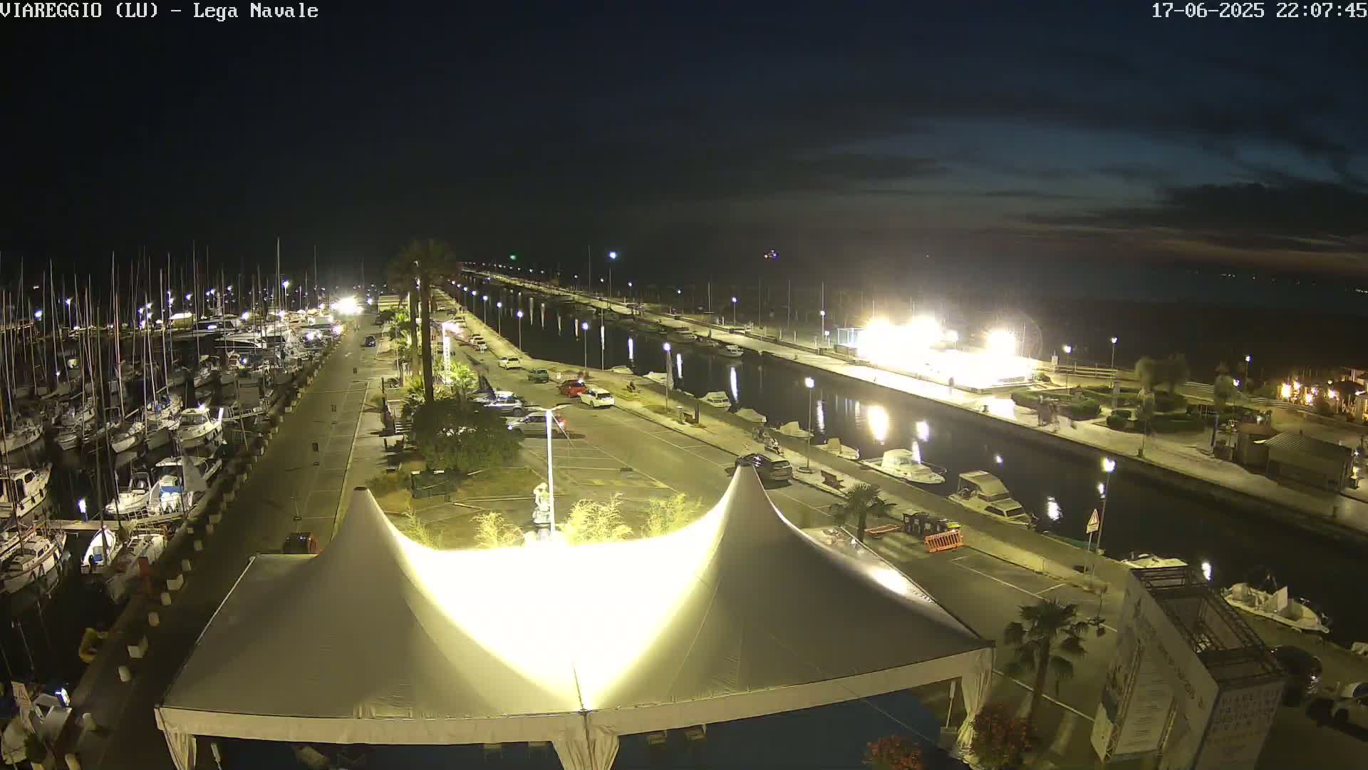 A nighttime view of a marina with numerous boats docked, a paved walkway lined with cars and streetlights, and a large white tent in the foreground, under a partly cloudy sky.