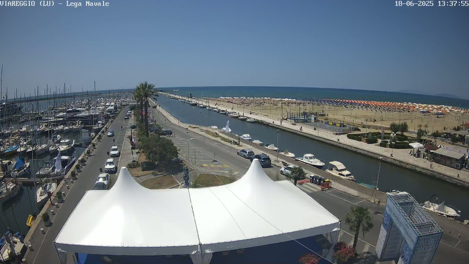 A sunny day shows a marina filled with boats, a canal separating it from a beach lined with umbrellas, and a roadway with cars leading to a white tent in the foreground.