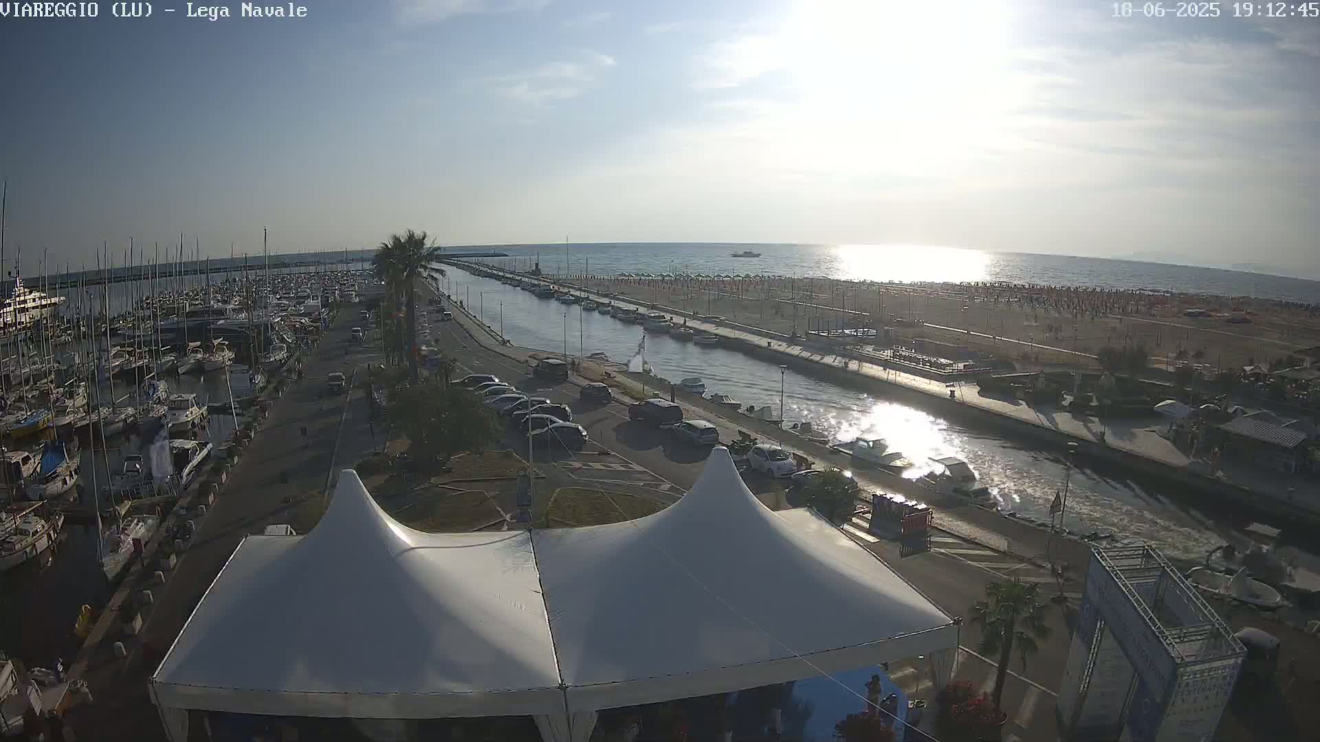 A marina filled with boats is next to a canal that leads to a beach with many people under a sunny sky.