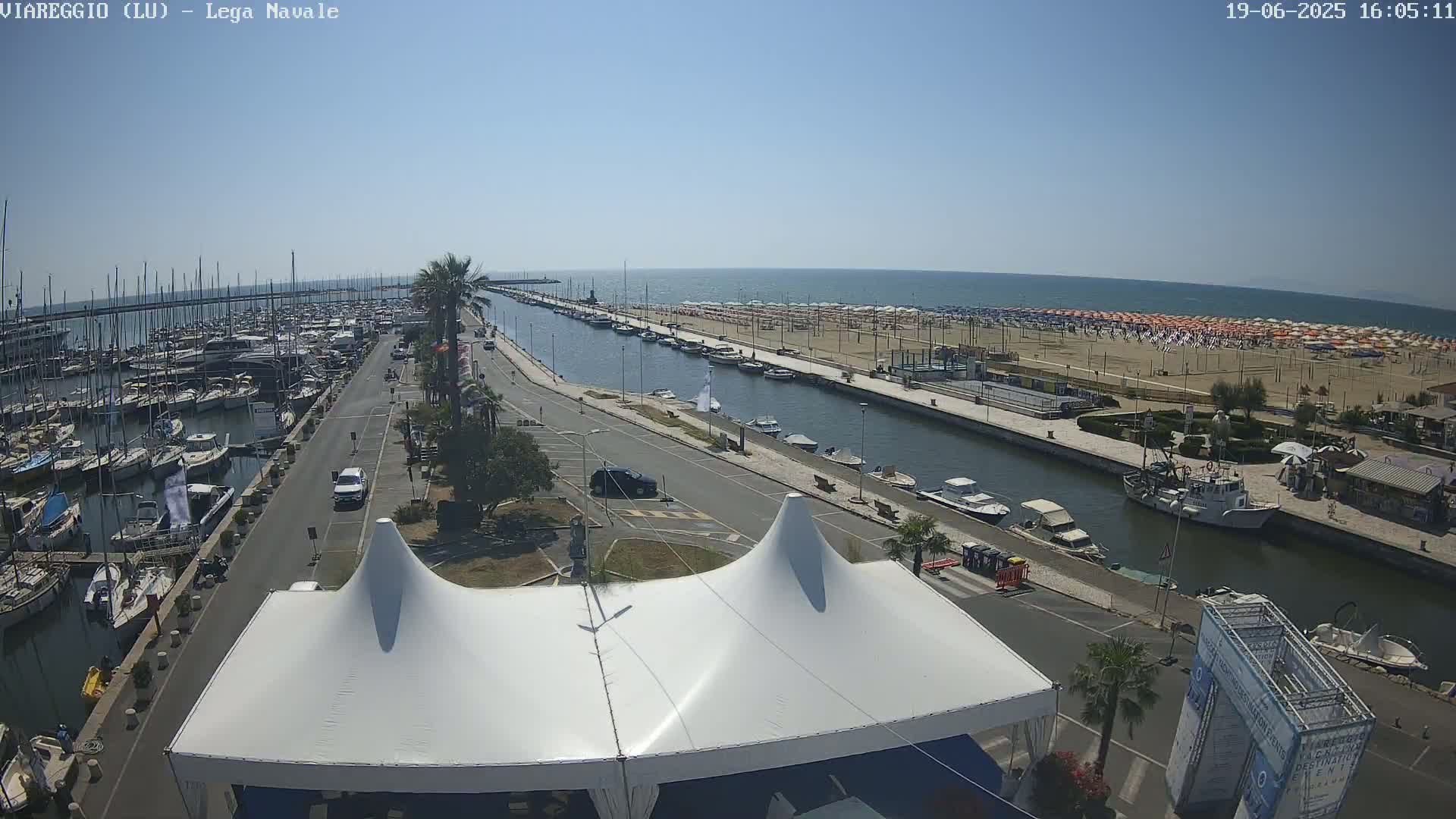 A marina filled with boats is next to a canal that leads to a beach with numerous umbrellas under a clear, sunny sky.