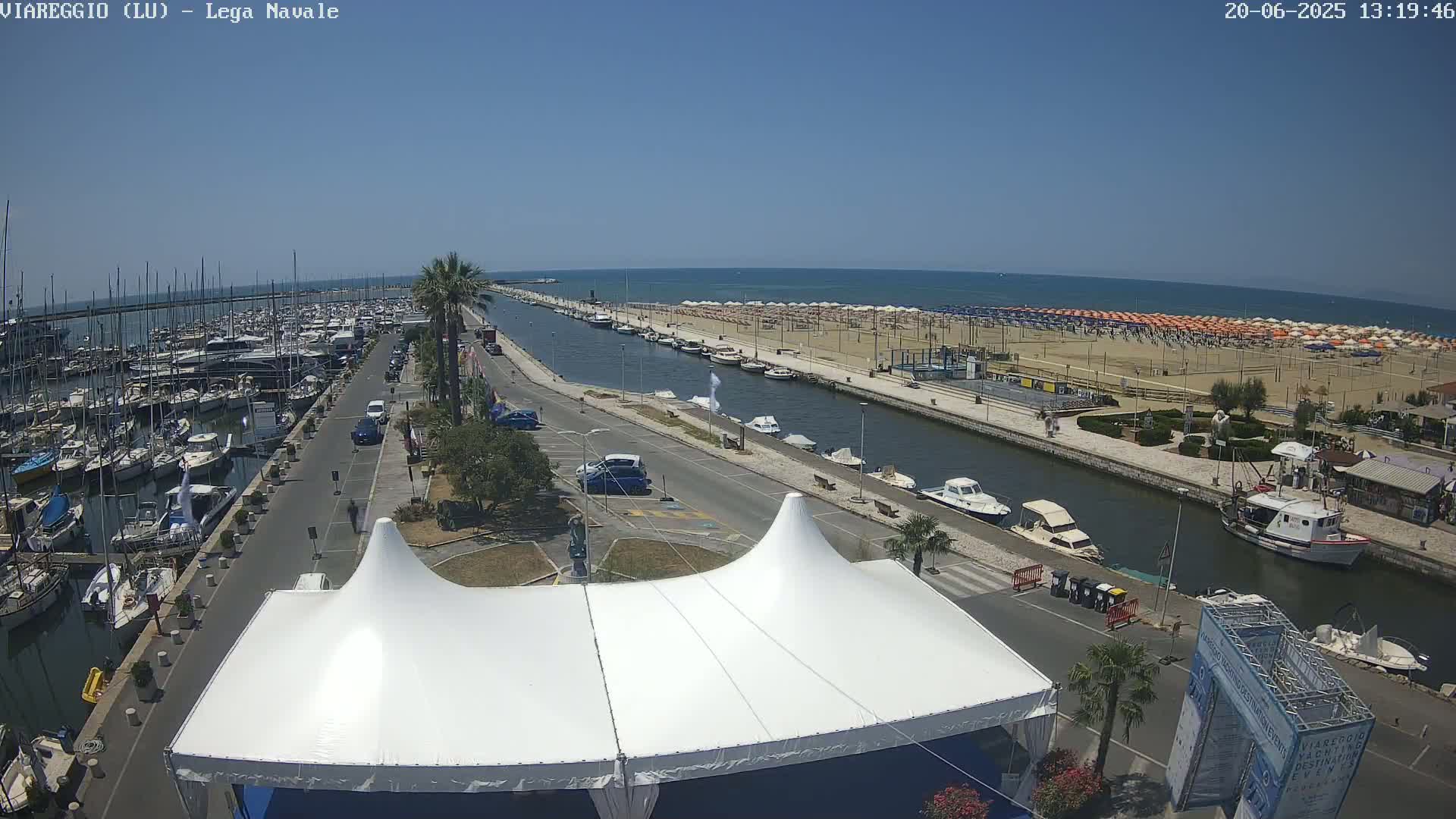A marina filled with boats, a canal, a sandy beach with numerous umbrellas, and a large white tent are visible under a clear blue sky.