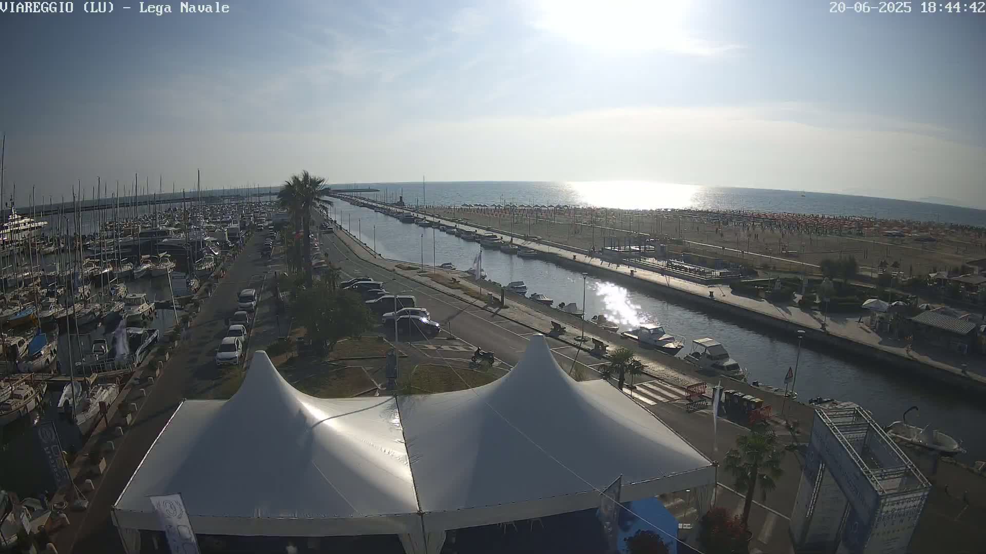 A sunny day at a marina shows a canal separating a crowded beach from a harbor full of boats, with two large white tents in the foreground.