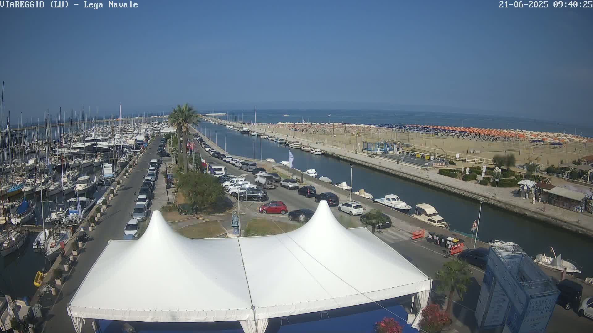 A marina filled with boats, a canal, a beach with numerous umbrellas, and a street lined with parked cars are visible on a sunny day.
