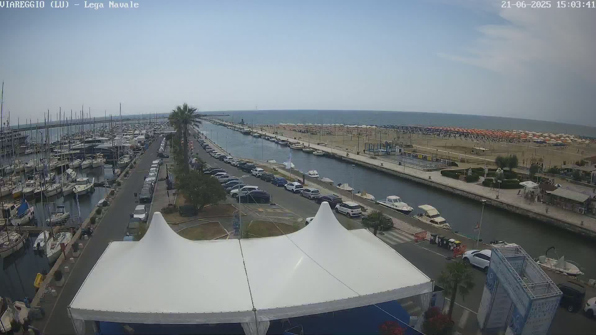 A marina filled with boats is next to a canal, which leads to a beach covered with orange umbrellas under a partly cloudy sky.