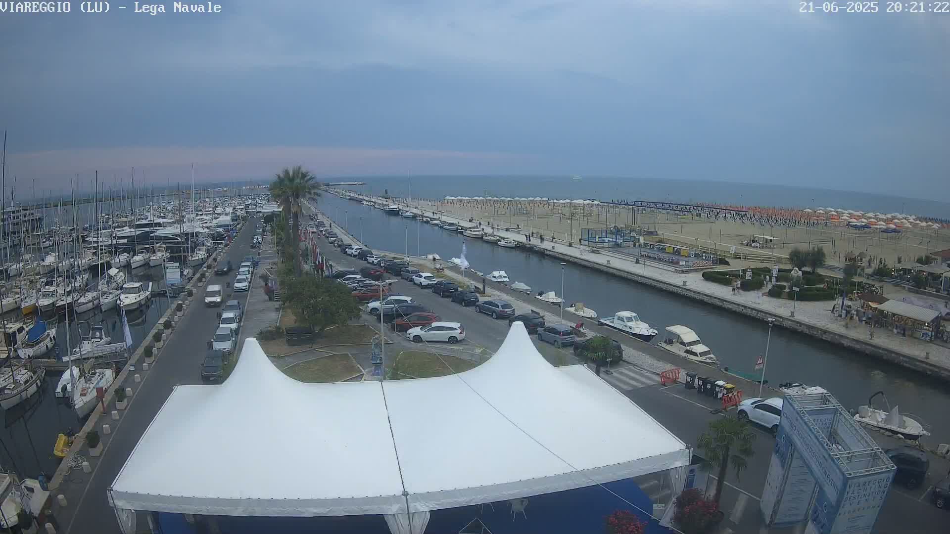 A marina filled with boats is adjacent to a canal that leads to a beach with numerous umbrellas under an overcast sky.