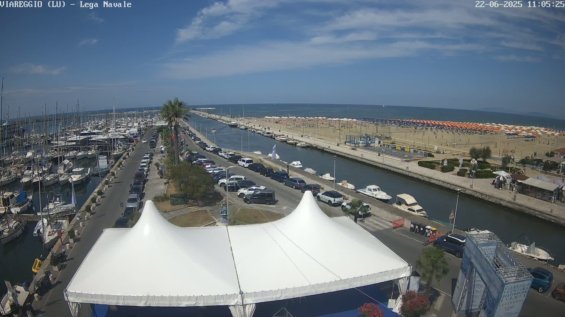 A marina filled with boats is adjacent to a canal, which runs alongside a beach crowded with people under numerous orange umbrellas on a sunny day.