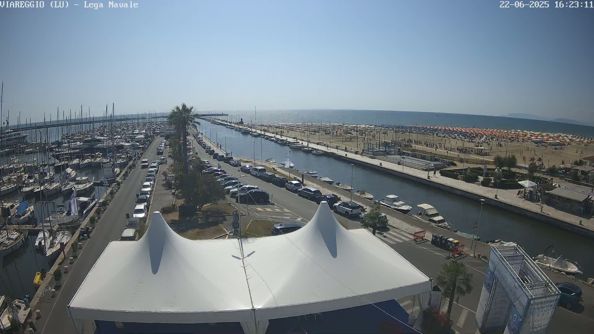 A harbor filled with boats, a canal lined with parked cars and beach umbrellas, and a sandy beach under a clear blue sky.