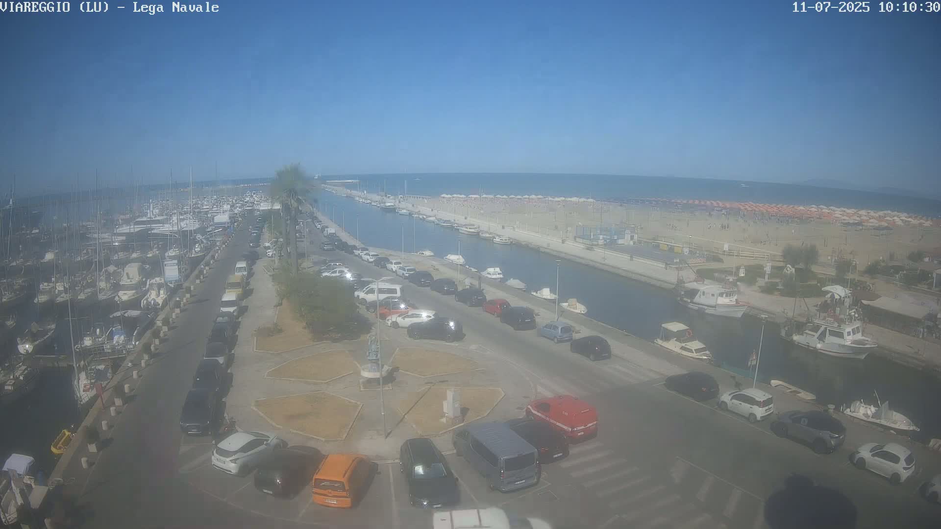 A harbor filled with boats is adjacent to a canal that leads to a crowded beach on a sunny day.