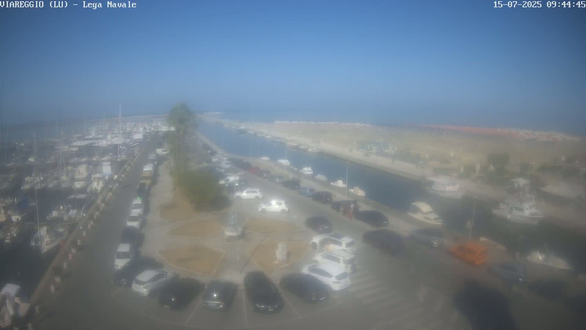 An aerial view shows a marina filled with boats, a canal, a parking lot with cars, and a hazy, sunny day.
