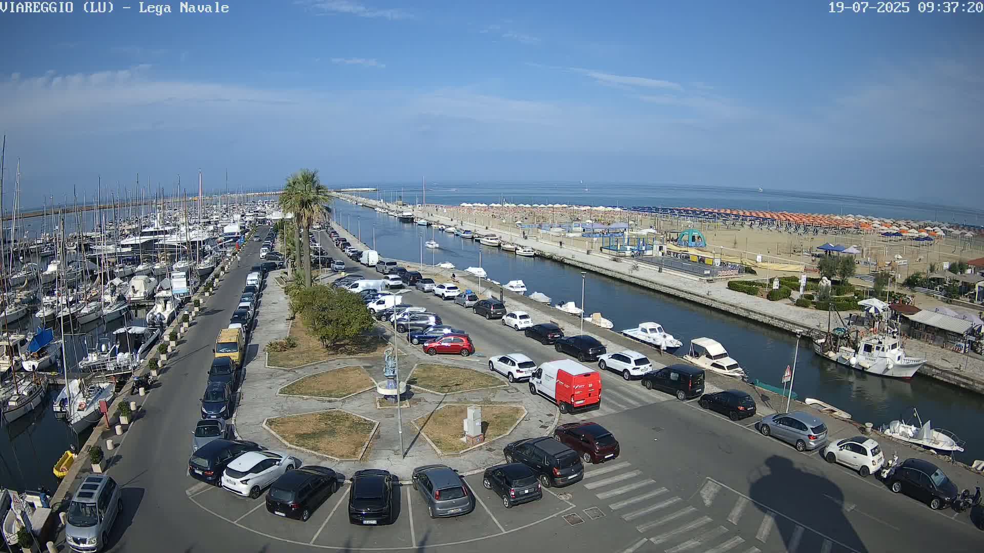 A marina filled with boats is adjacent to a parking lot and a canal leading to a beach with many umbrellas under a clear, sunny sky.