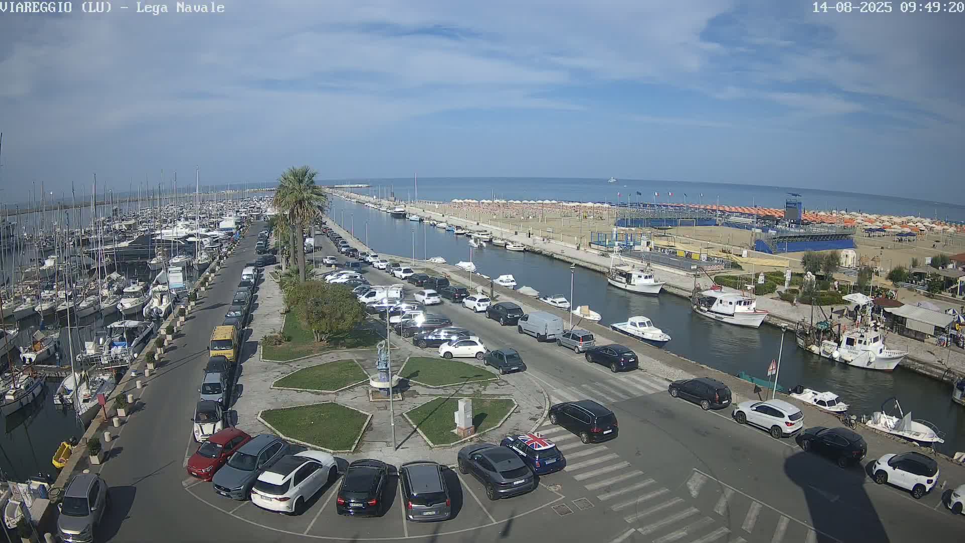 A harbor filled with boats is next to a canal lined with parked cars and a beach under a mostly sunny sky.