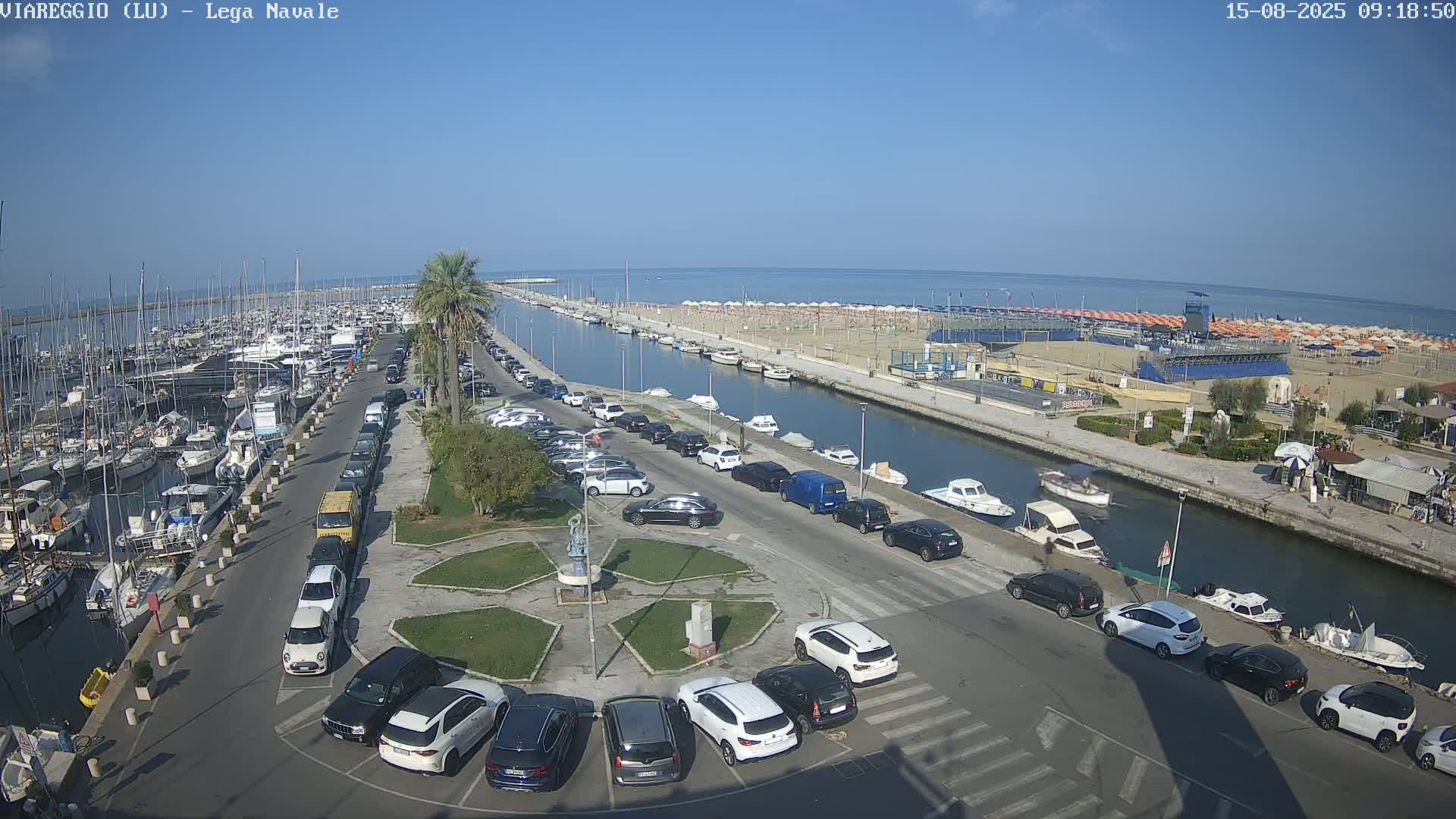 A harbor filled with boats is adjacent to a canal lined with parked cars and a sandy beach under a clear sunny sky.