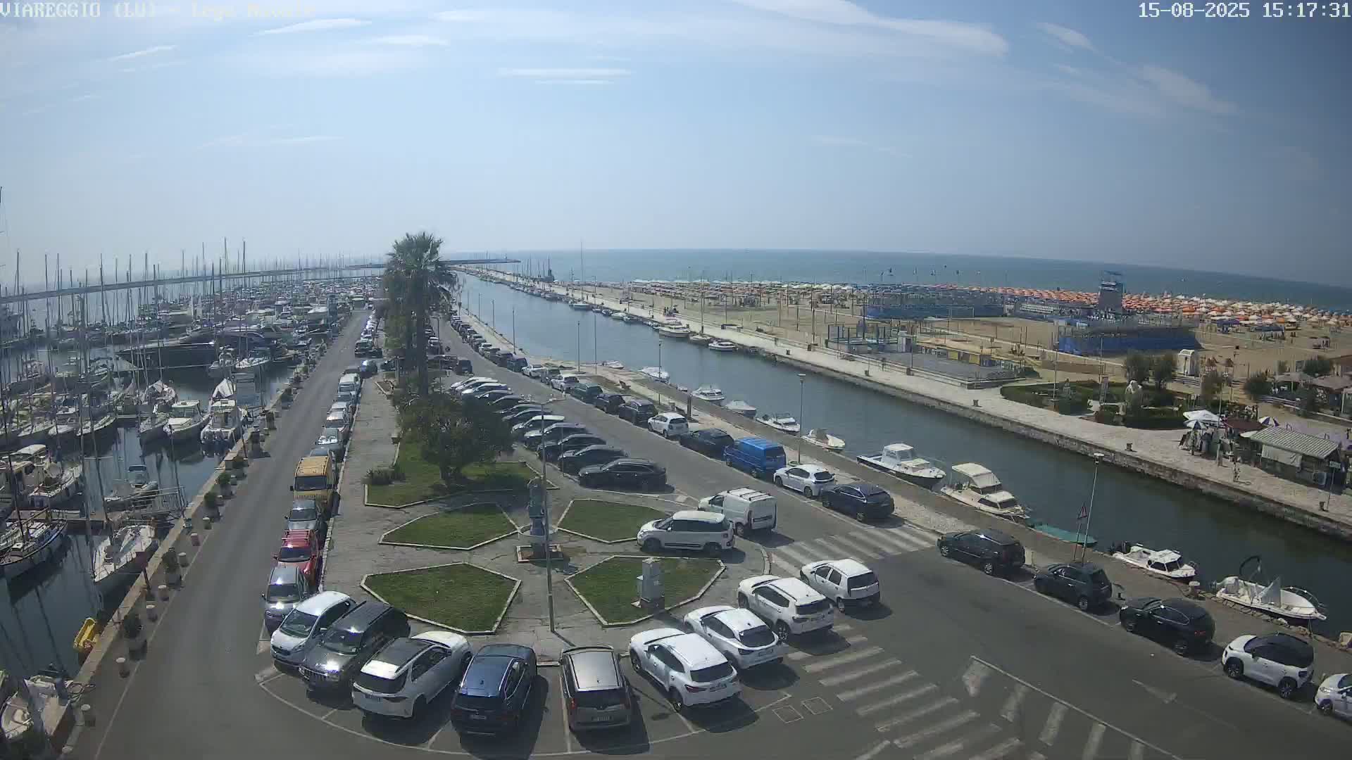 A harbor filled with boats is adjacent to a canal lined with parked cars and a sandy beach under a clear sunny sky.