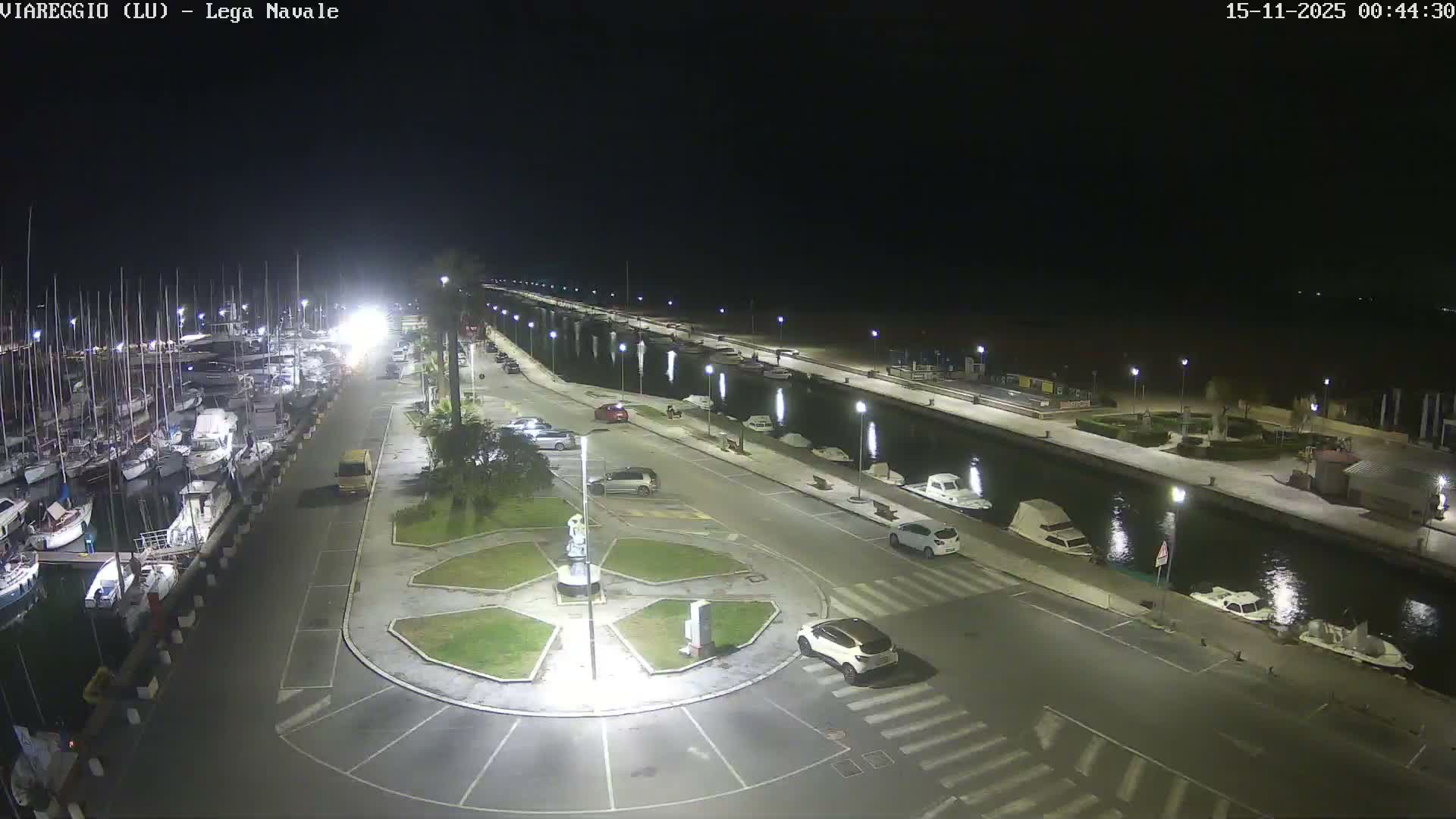 A clear night reveals a brightly illuminated marina or canal system, featuring numerous boats docked along waterways, a central road with a roundabout and vehicles, and streetlights reflecting across the calm water that extends into the distance.
