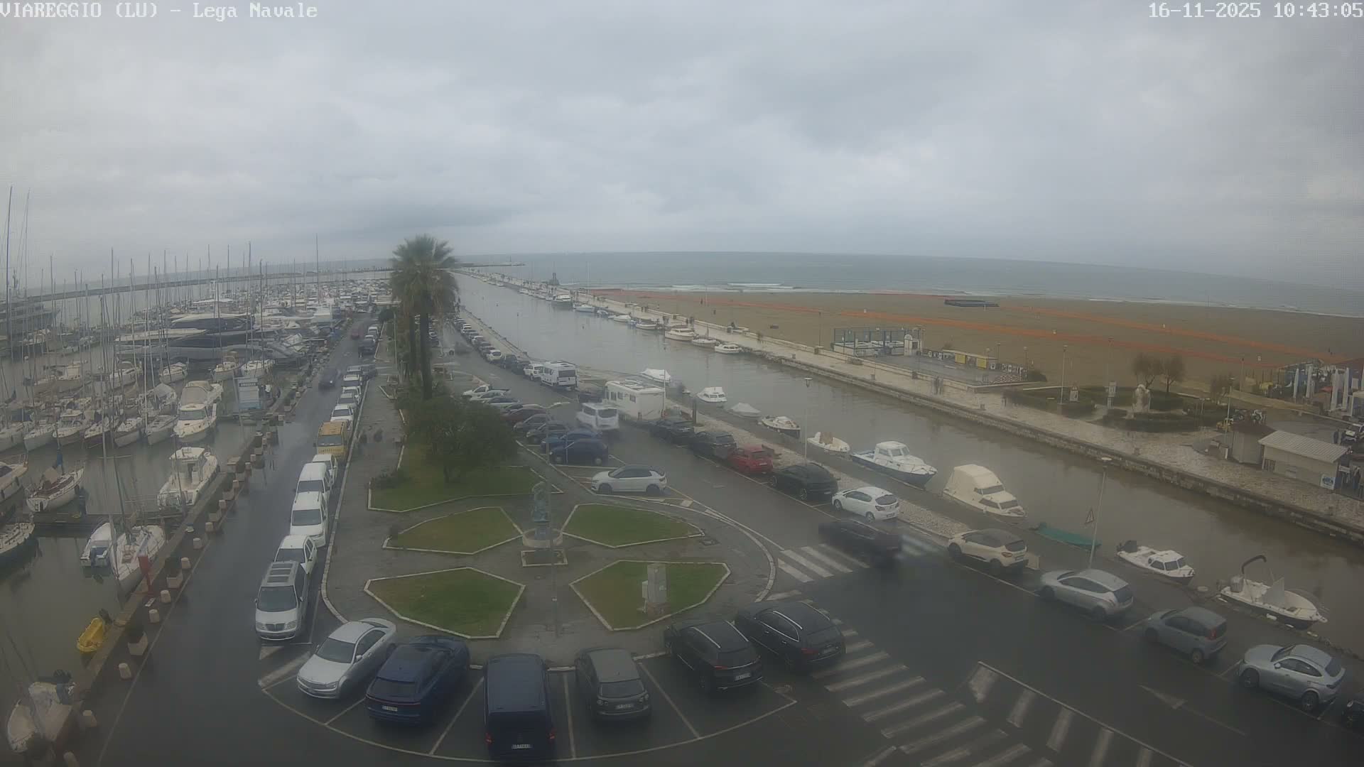 An elevated view captures a busy marina filled with boats and surrounding roads with parked cars, a canal leading to a wide sandy beach beside the ocean, all on a dim, overcast day with wet ground suggesting rain.