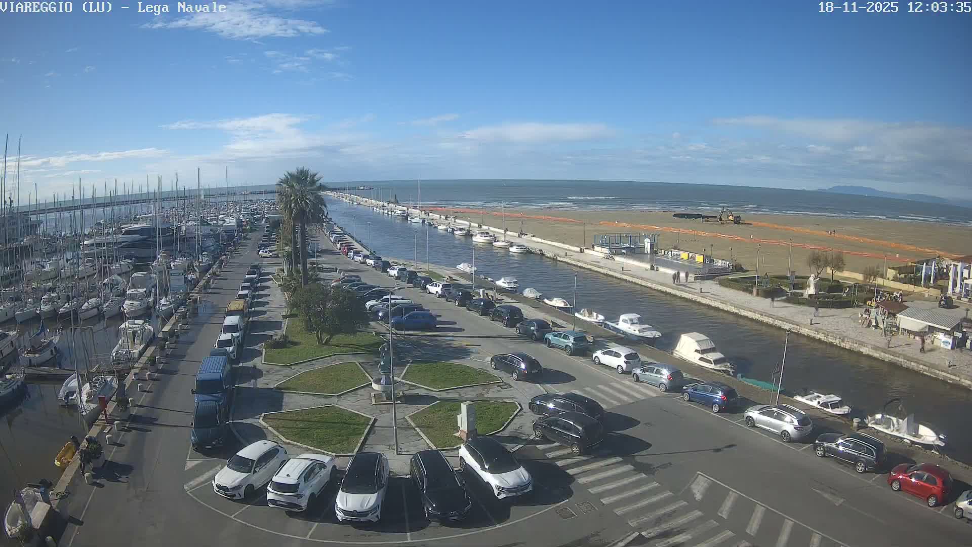An elevated view captures a busy marina filled with boats and surrounding roads with parked cars, a canal leading to a wide sandy beach beside the ocean, all on a dim, overcast day with wet ground suggesting rain.