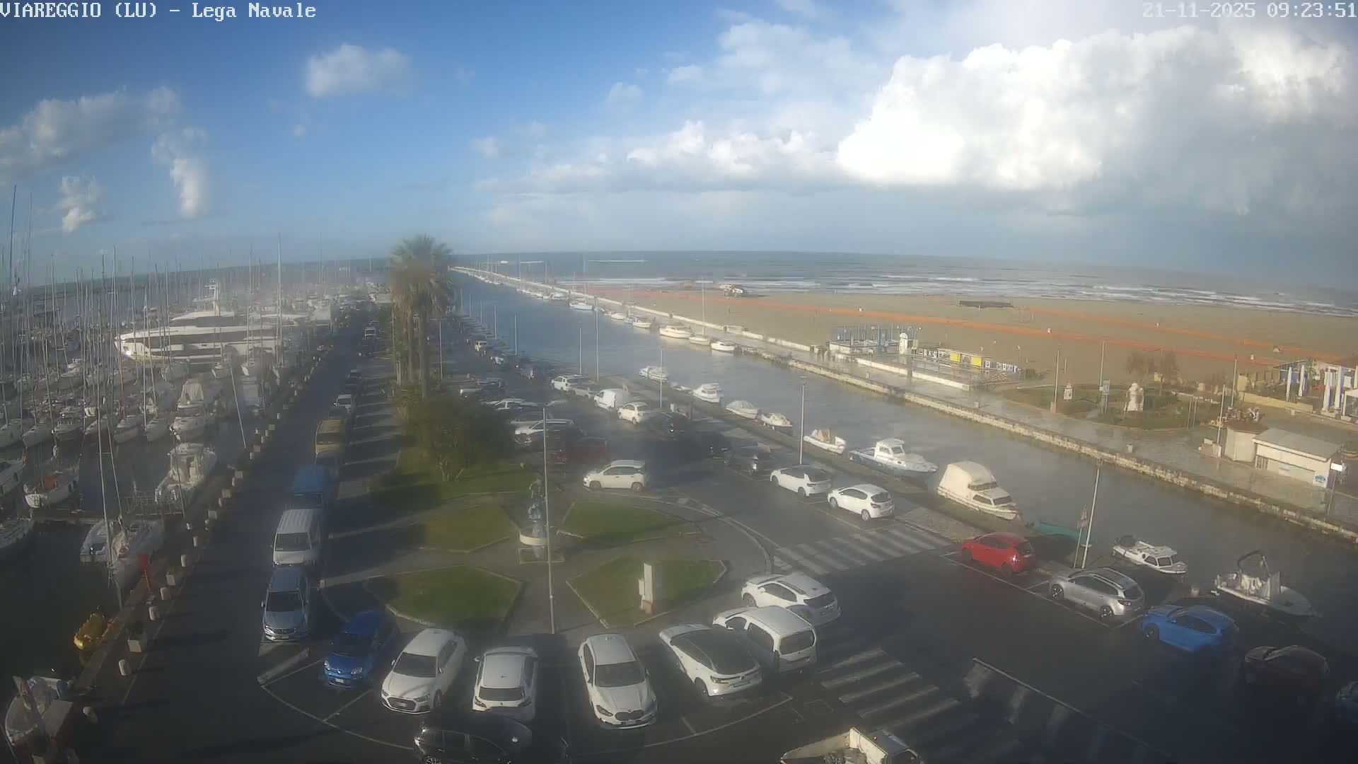 An elevated view captures a marina crowded with sailboats and yachts, a canal lined with moored small boats and a road full of parked cars, extending towards a wide sandy beach and the sea under a partly cloudy sky.