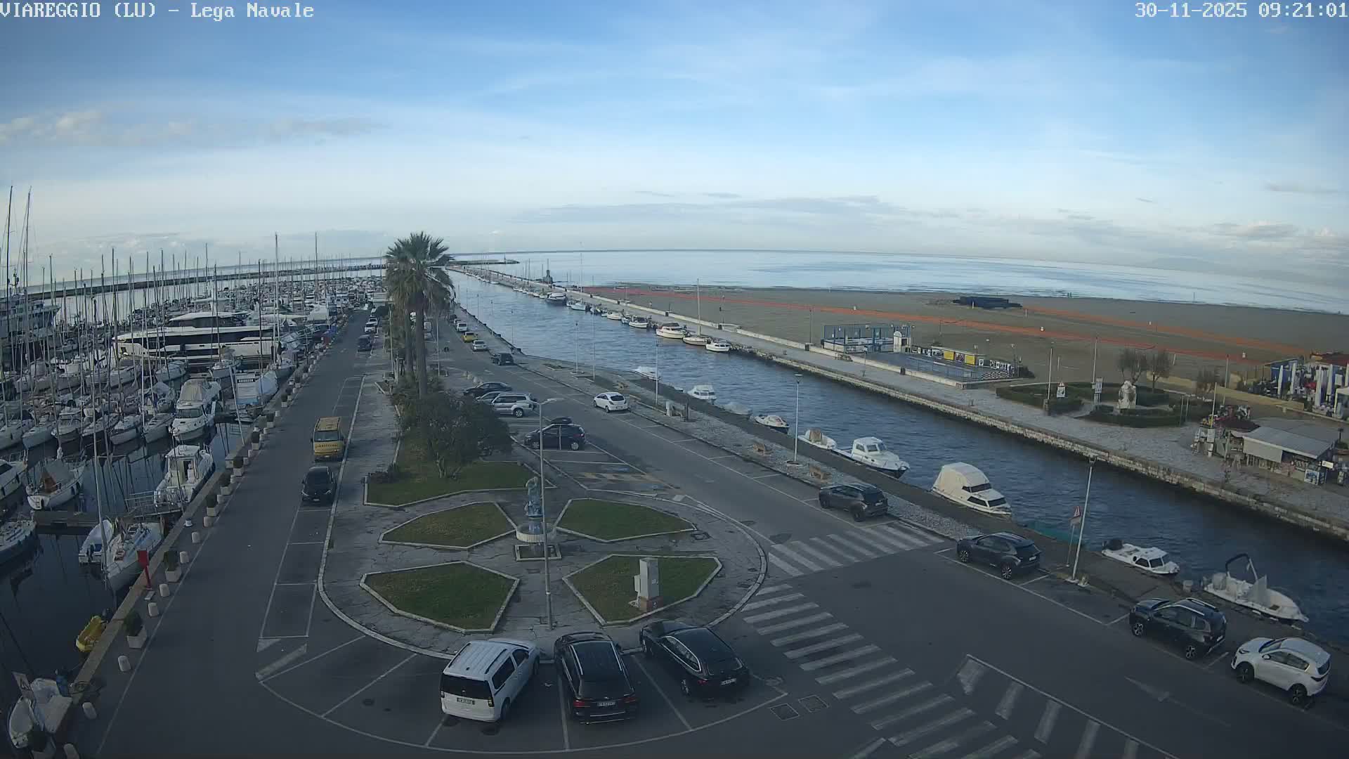 A coastal landscape features a bustling marina filled with boats, a canal lined with roads, cars, and palm trees extending to a wide sandy beach and the ocean, all beneath a bright, partly cloudy sky.