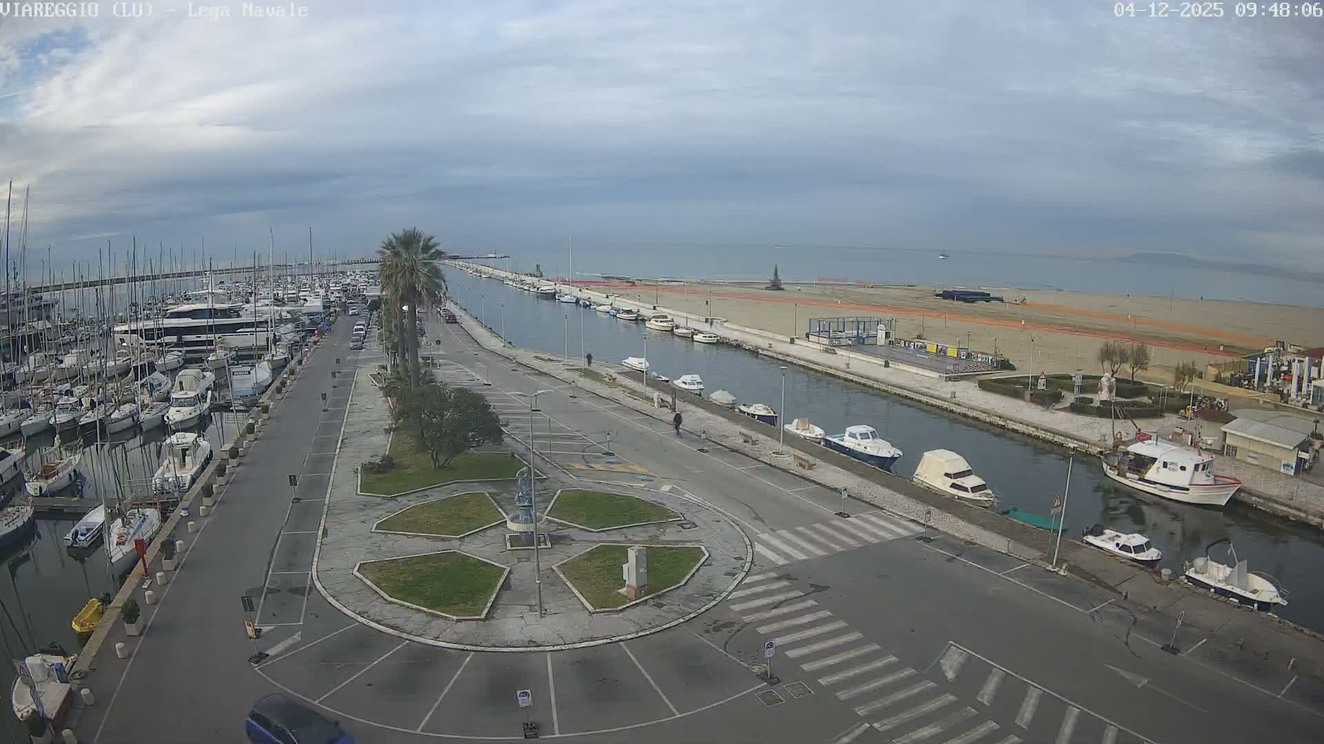 Under a cloudy sky, a wide panorama captures a bustling marina filled with numerous sailboats and yachts on the left, a parallel canal with smaller boats, a road, a sandy beach, and the calm sea stretching to the horizon on the right.
