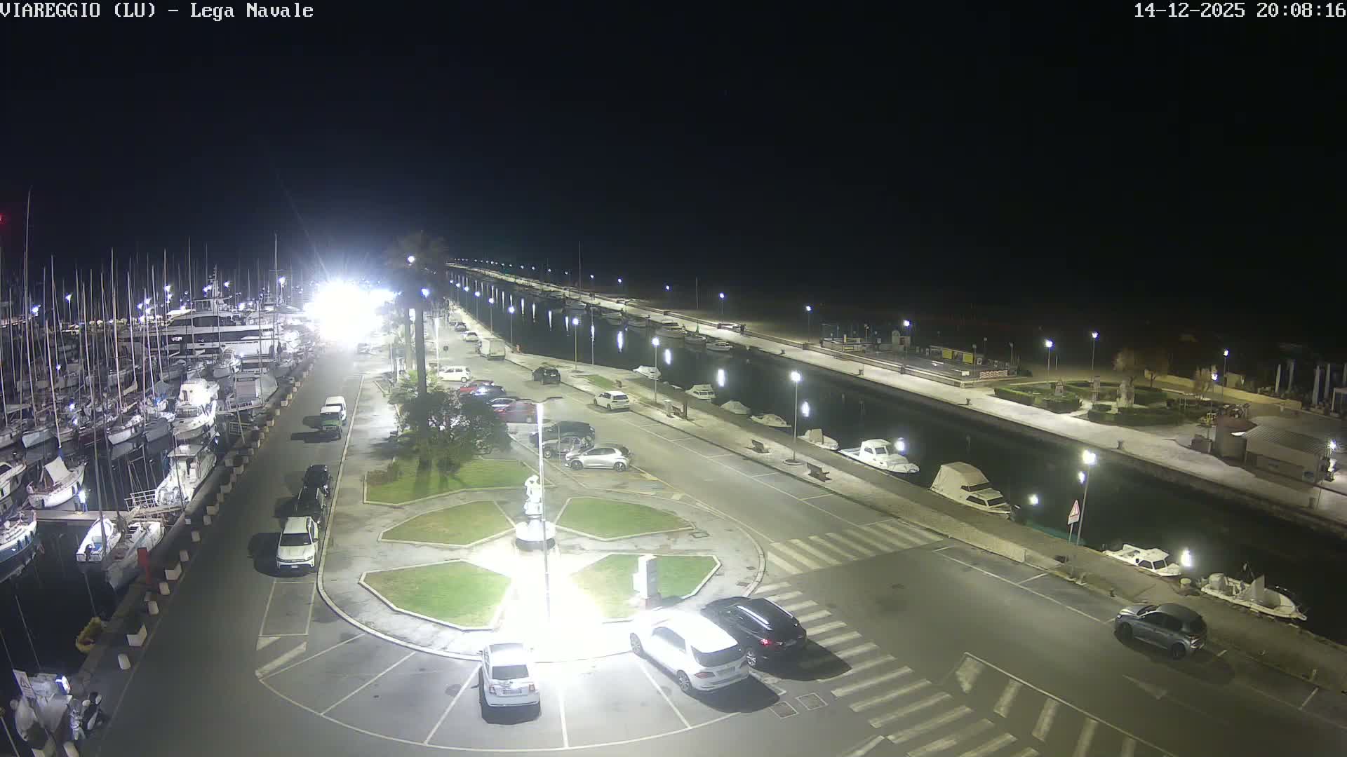 Under a cloudy sky, a wide panorama captures a bustling marina filled with numerous sailboats and yachts on the left, a parallel canal with smaller boats, a road, a sandy beach, and the calm sea stretching to the horizon on the right.